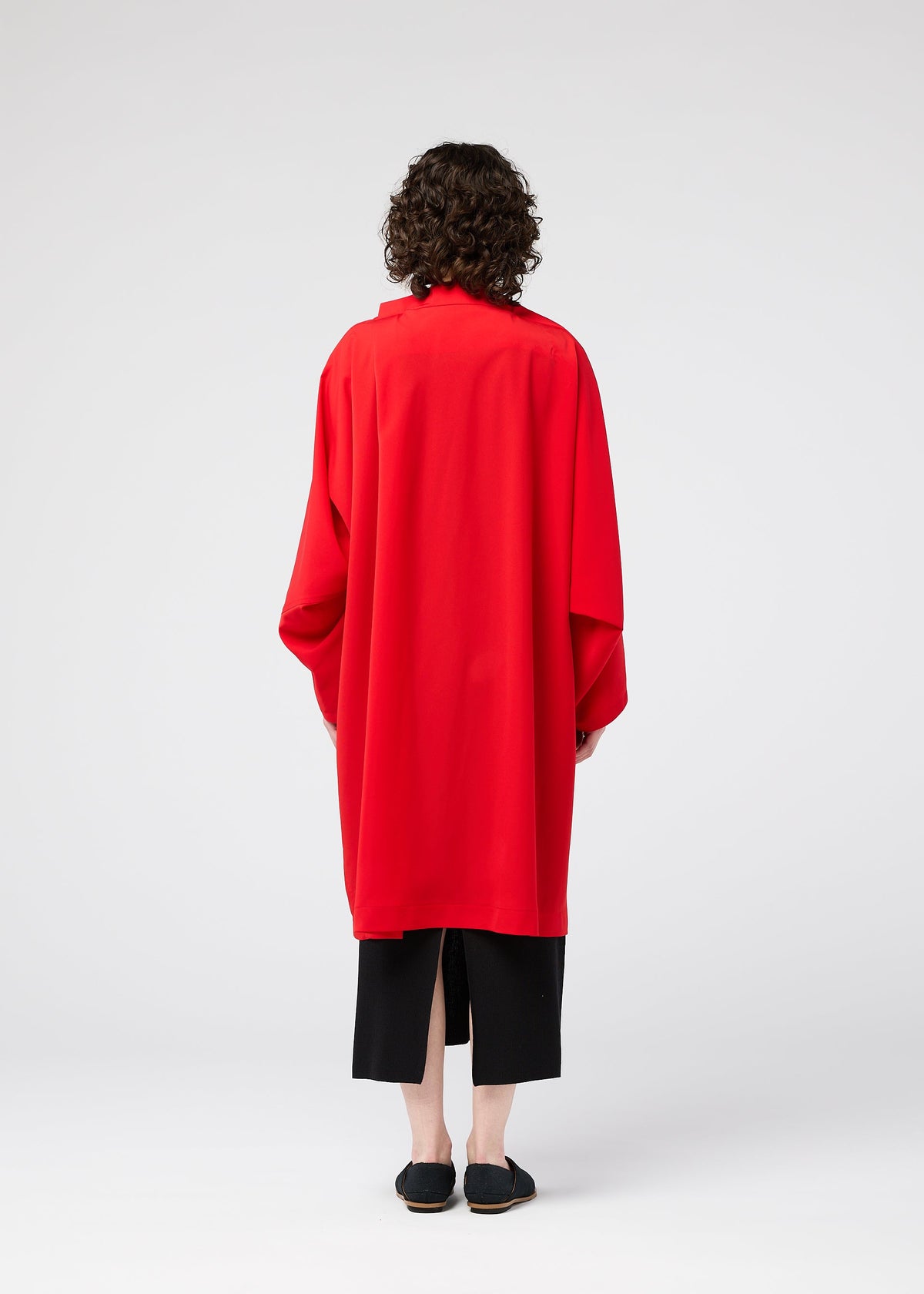 A person with curly hair faces away, wearing the PARALLEL WRAP by 132 5. ISSEY MIYAKE—a loose, bright red lightweight coat—over wide black cropped pants and black shoes, set against a plain white background. view 8 of 11