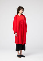 A person with curly hair wears the PARALLEL WRAP by 132 5. ISSEY MIYAKE, a loose-fitting bright red coat, over black cropped pants and black shoes, standing against a plain white background.