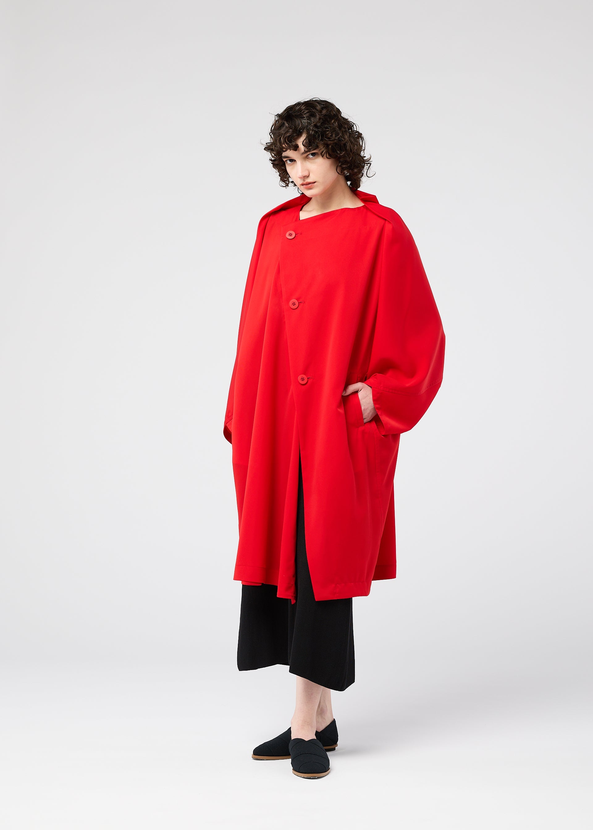 Zoomed view A person with curly hair stands against a white background, wearing the PARALLEL WRAP coat by 132 5. ISSEY MIYAKE—bright red, wrinkle-resistant, large buttons—paired with black wide-leg pants and slip-on shoes, hands in pockets.