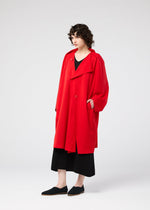 A person with curly hair stands against a plain background, wearing the PARALLEL WRAP by 132 5. ISSEY MIYAKE—a long, loose-fitting lightweight red coat—over black wide-legged pants and black shoes.