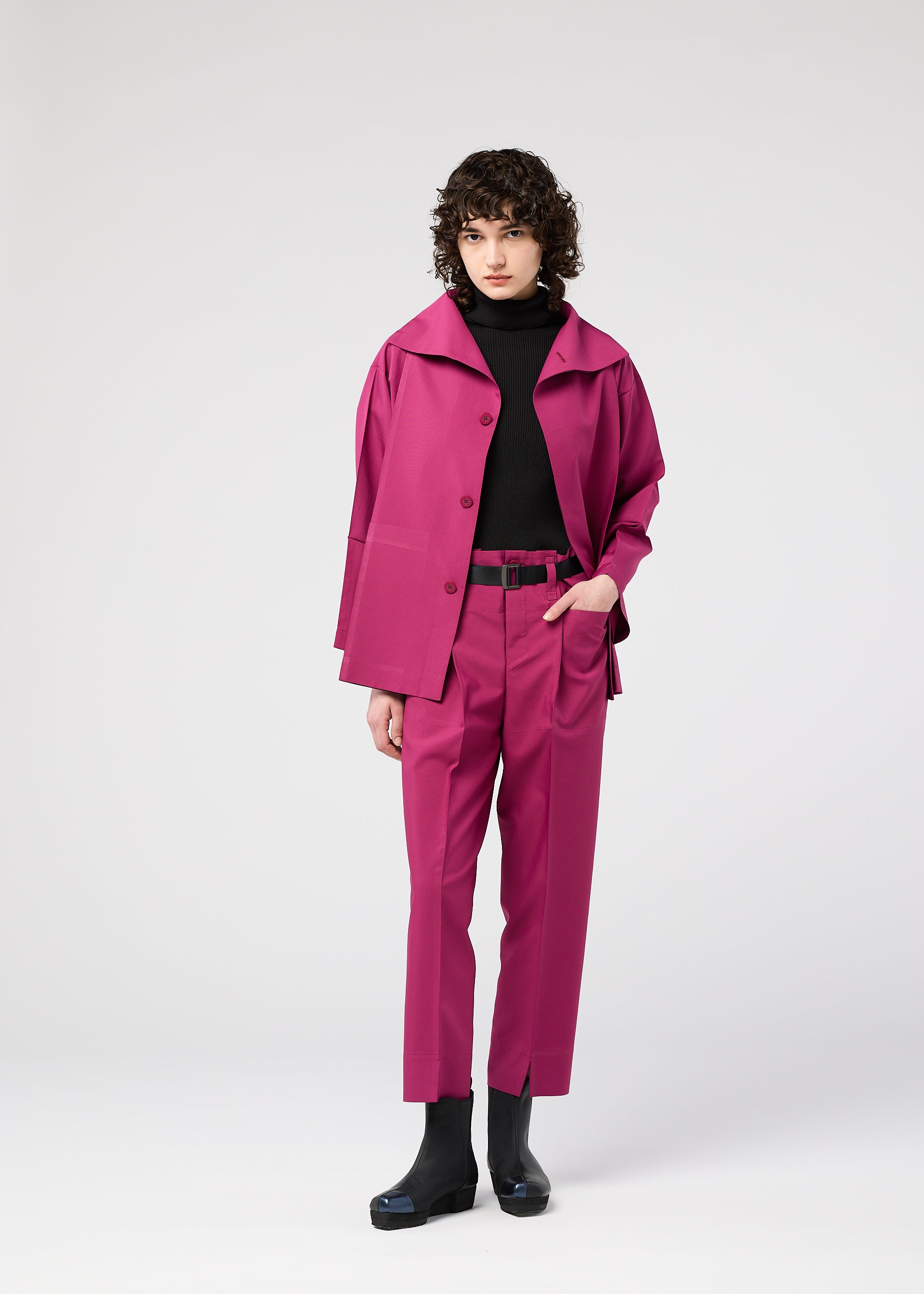 Zoomed view A person with curly hair wears the magenta 132 5. FLAT 1 suit by 132 5. ISSEY MIYAKE—matching pants and jacket, black turtleneck, belt, and boots—standing against a plain light gray background.
