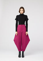 A woman stands against a plain, light background, wearing a black turtleneck sweater and the VERTEX BOTTOMS by 132 5. ISSEY MIYAKE—bright magenta pants with angular hips—paired with black ankle boots and a neutral expression.