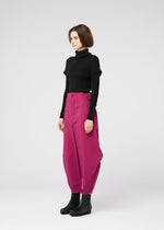 A woman with short brown hair and a neutral expression wears black ankle boots, a black turtleneck, and VERTEX BOTTOMS by 132 5. ISSEY MIYAKE—bright magenta 3D pants with unique draping—against a plain light background.