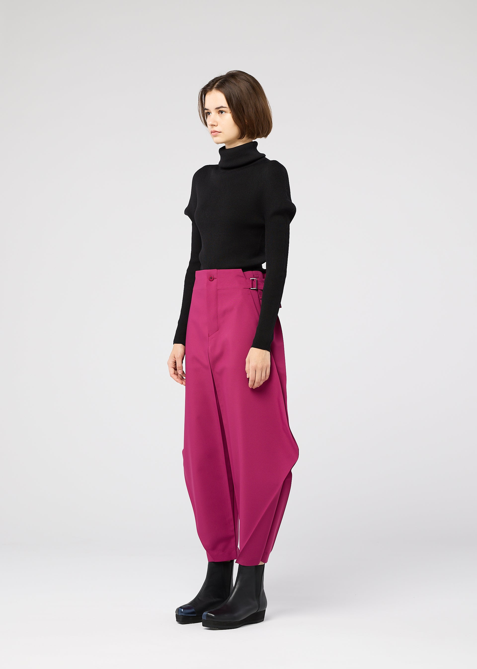 Zoomed view A woman with short brown hair and a neutral expression wears black ankle boots, a black turtleneck, and VERTEX BOTTOMS by 132 5. ISSEY MIYAKE—bright magenta 3D pants with unique draping—against a plain light background.