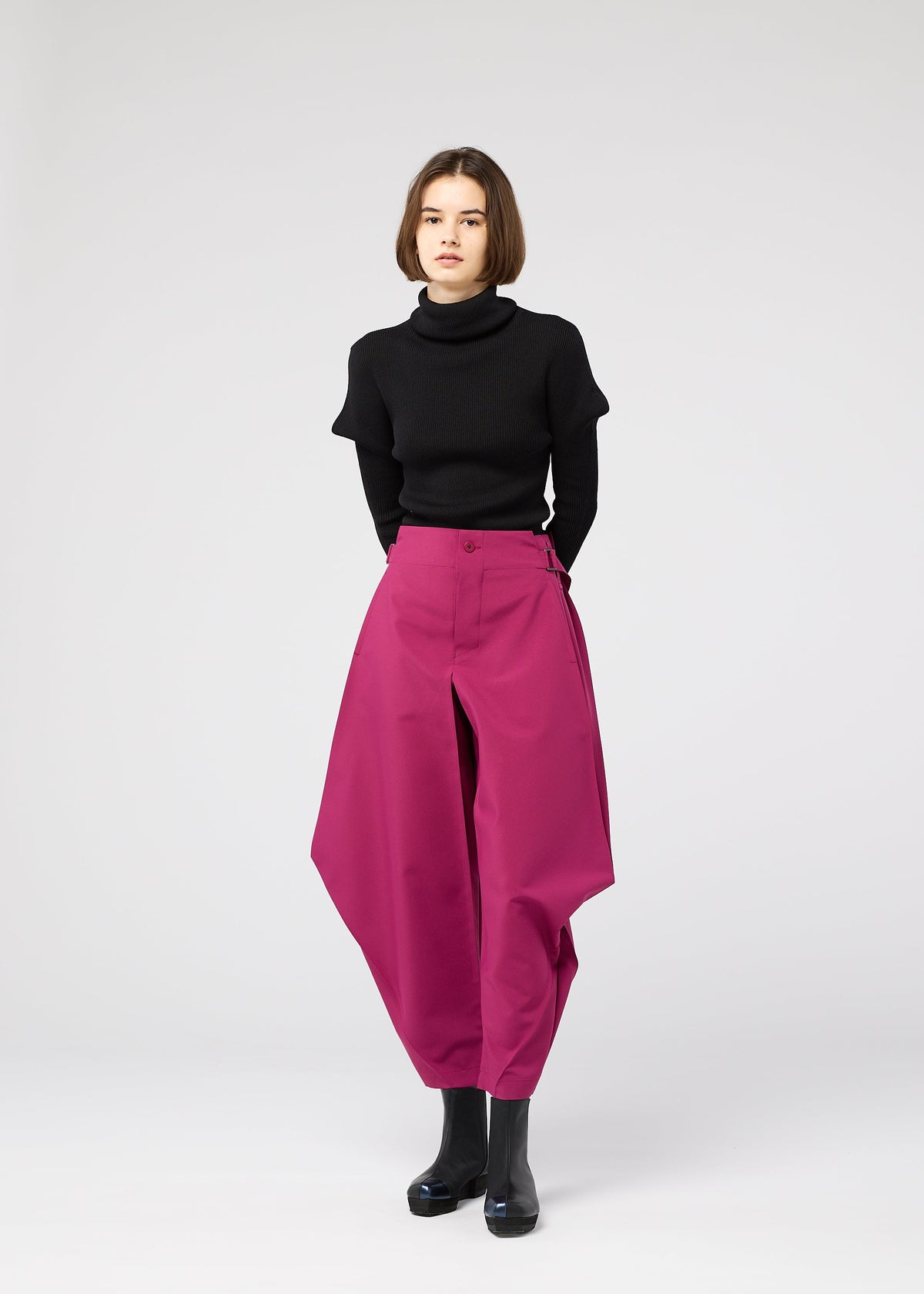 A woman with short brown hair stands against a white background, wearing a black turtleneck and the VERTEX BOTTOMS by 132 5. ISSEY MIYAKE—magenta pants with angular, three-dimensional design—paired with black ankle boots. view 11 of 12