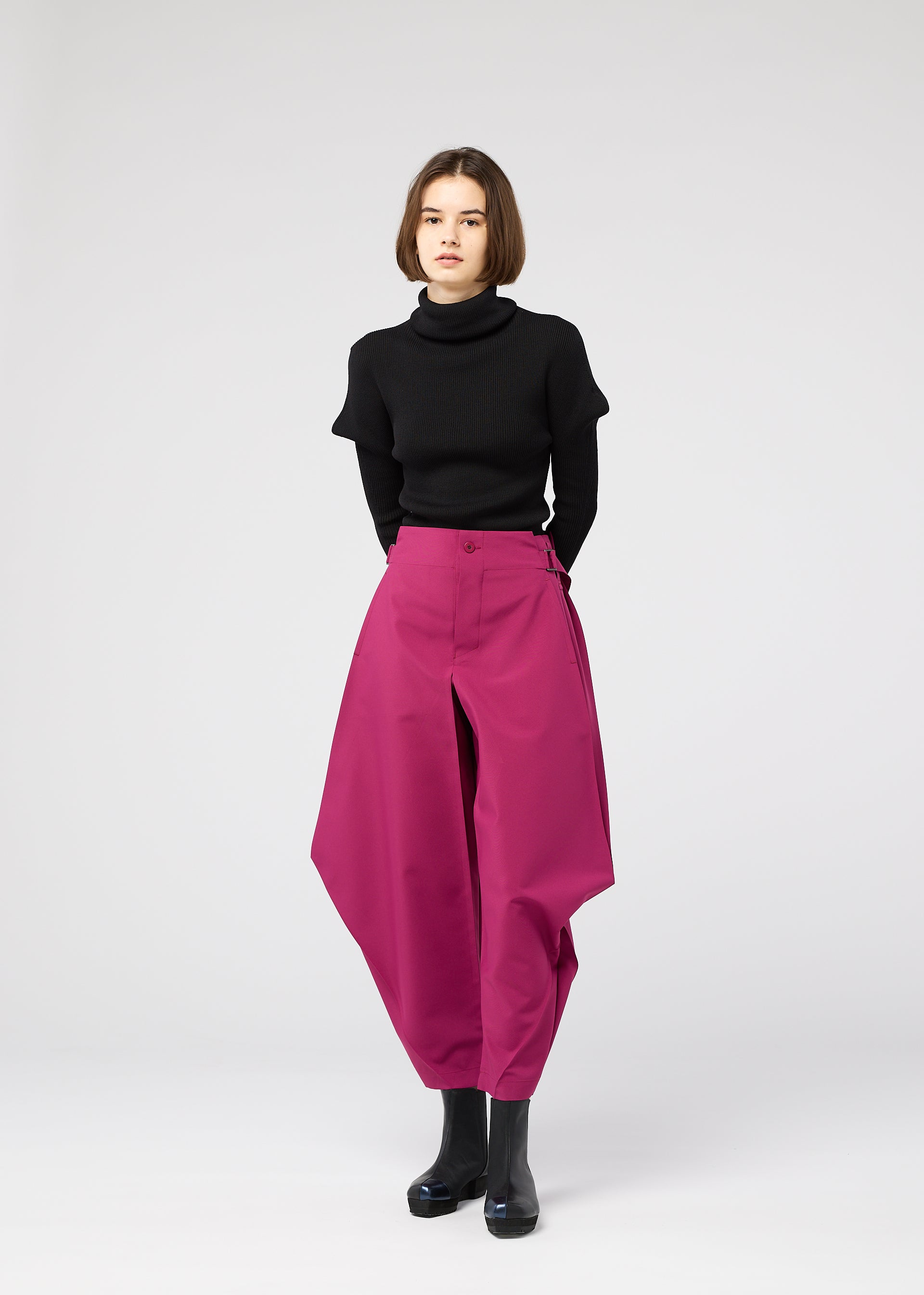 Zoomed view A woman with short brown hair stands against a white background, wearing a black turtleneck and the VERTEX BOTTOMS by 132 5. ISSEY MIYAKE—magenta pants with angular, three-dimensional design—paired with black ankle boots.