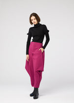 A woman with a bob wears a fitted black turtleneck, black ankle boots, and the VERTEX BOTTOMS by 132 5. ISSEY MIYAKE—voluminous magenta pants with an asymmetrical design—standing against a plain light background.