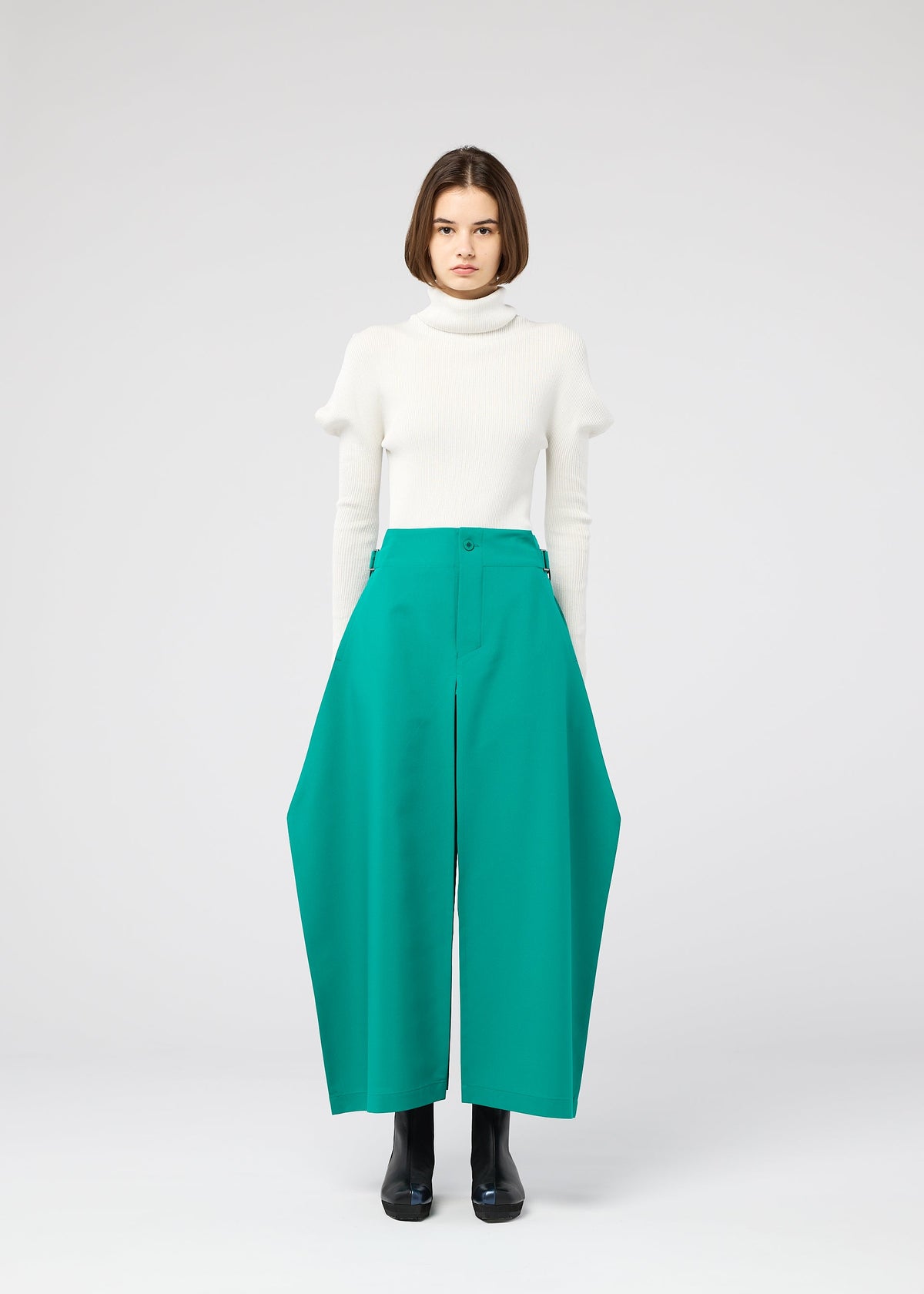 A woman with straight brown hair wears a white turtleneck sweater and the VERTEX BOTTOMS by 132 5. ISSEY MIYAKE—wide, three-dimensional green pants—paired with black shoes, standing against a plain light gray background. view 8 of 11