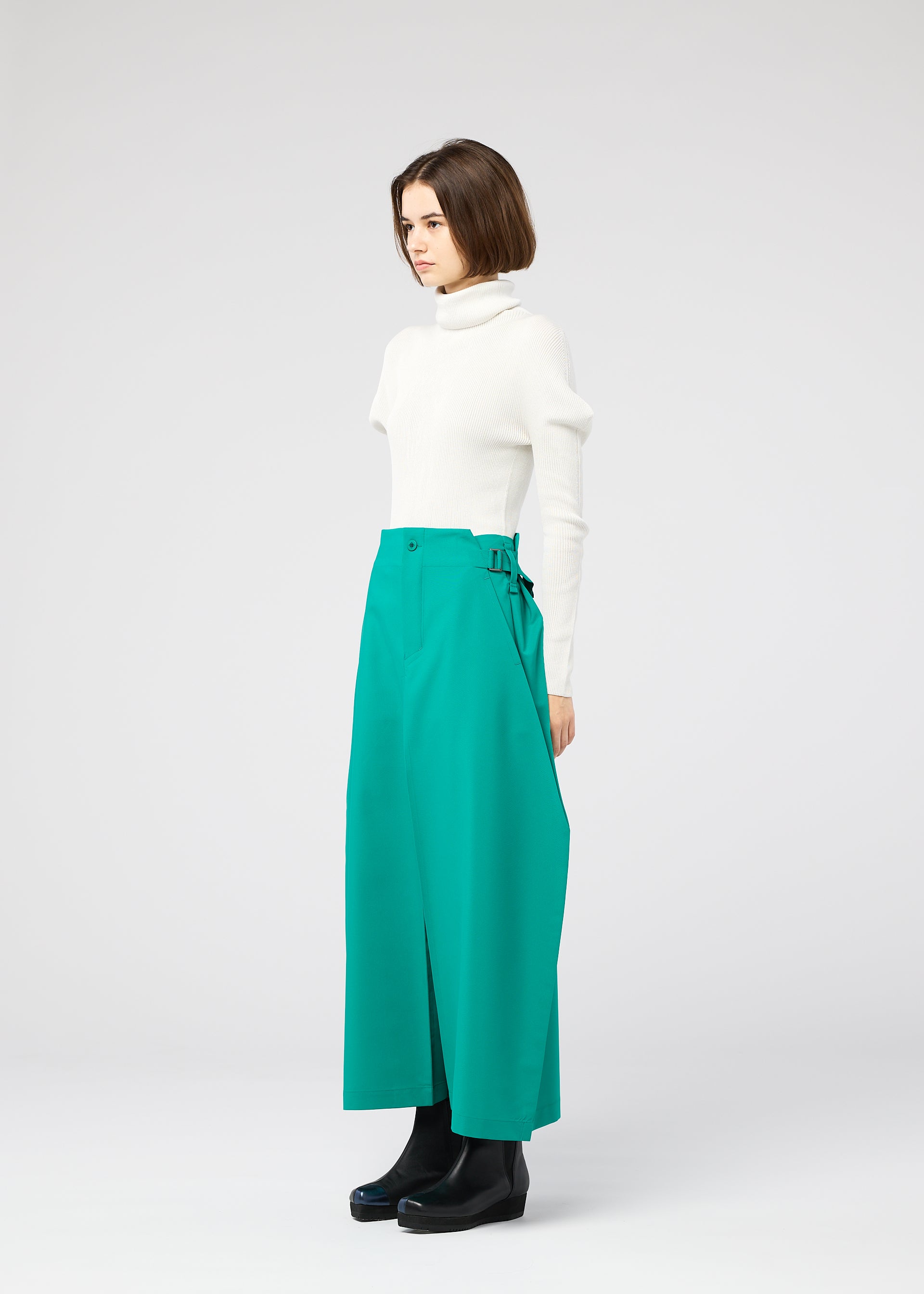 Zoomed view A woman with a bob haircut stands sideways in a white turtleneck, wearing 132 5. ISSEY MIYAKE VERTEX BOTTOMS—voluminous bright green pants—and black ankle boots against a plain light gray background.