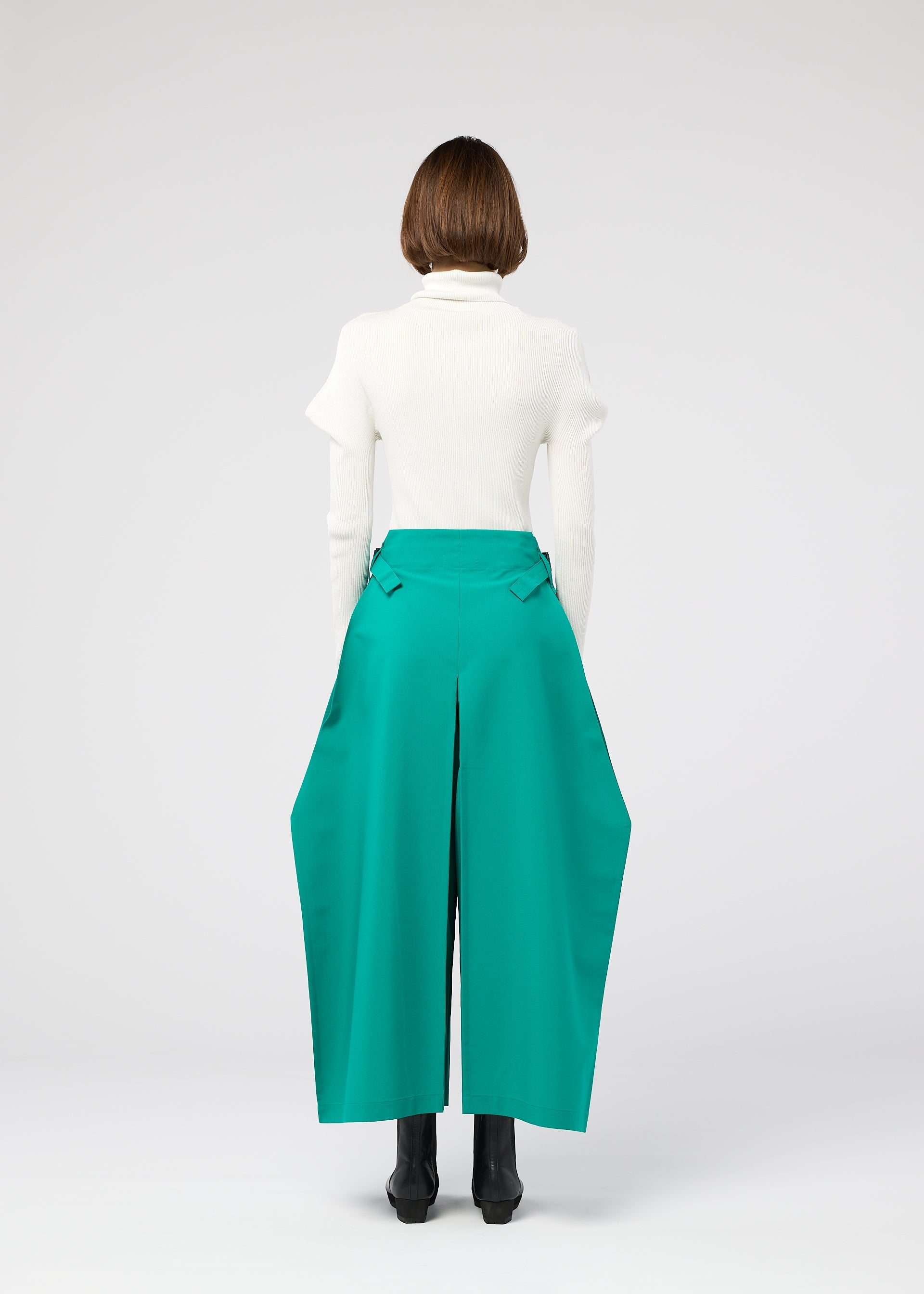 Zoomed view A person faces away, wearing a white turtleneck and the 132 5. ISSEY MIYAKE VERTEX BOTTOMS—voluminous teal green pants with angular panels—paired with black boots, set against a plain light gray background.