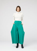 A woman with a bob haircut wears a white turtleneck sweater and the 132 5. ISSEY MIYAKE VERTEX BOTTOMS, posing with hands in pockets against a plain light gray background.