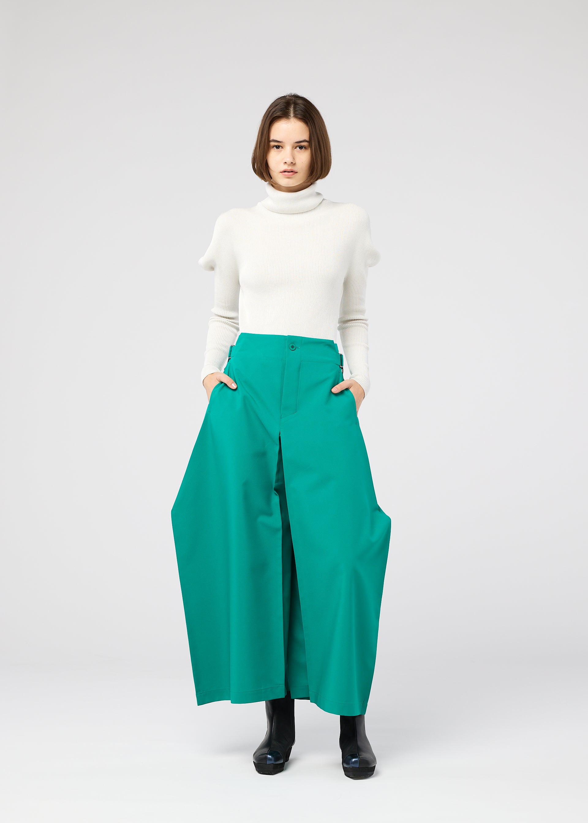 Zoomed view A woman with a bob haircut wears a white turtleneck sweater and the 132 5. ISSEY MIYAKE VERTEX BOTTOMS, posing with hands in pockets against a plain light gray background.