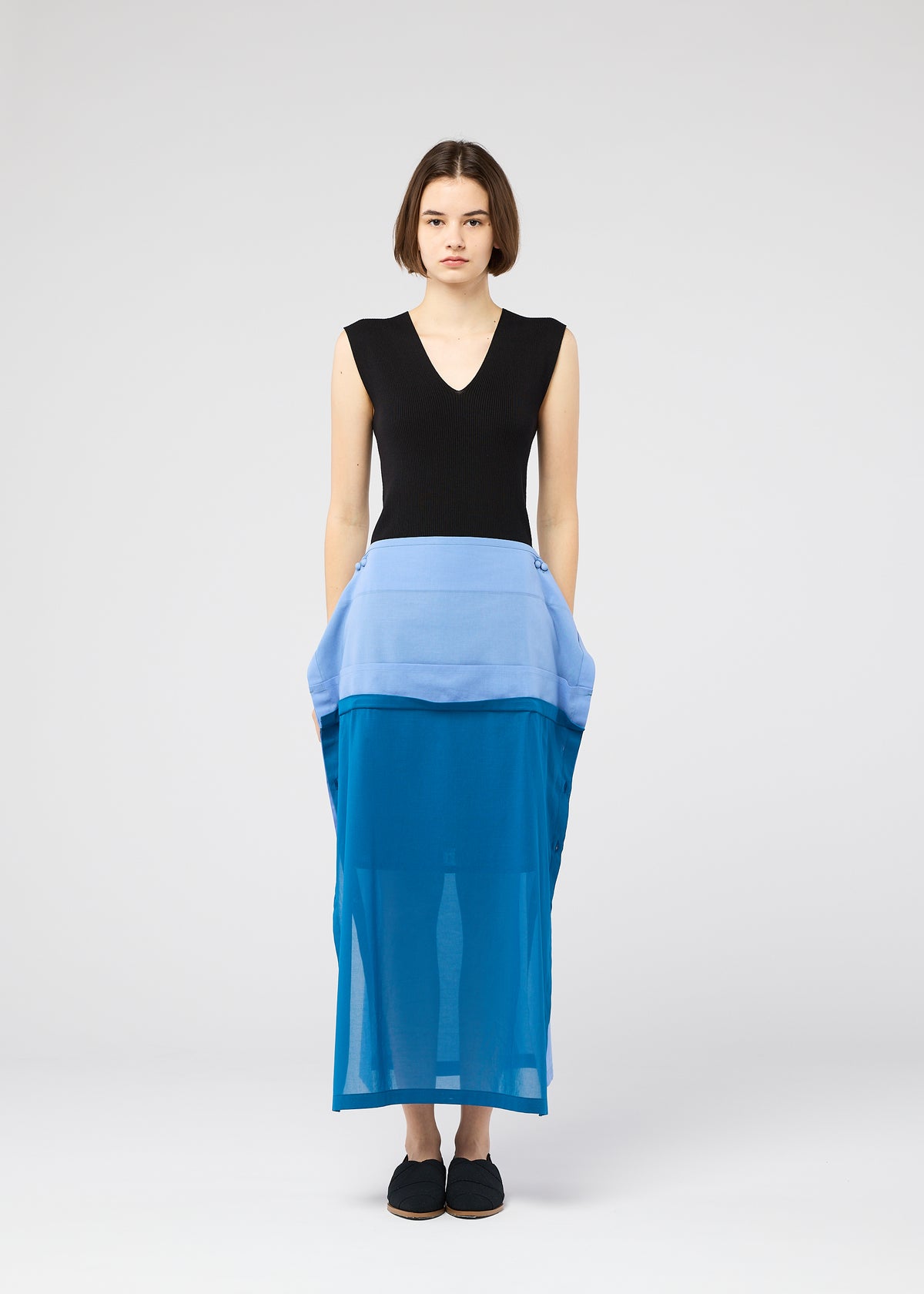 A woman with a short bob stands facing forward in the SHEER FOLIO sheer blue layered skirt by 132 5. ISSEY MIYAKE, paired with a sleeveless black V-neck top, posing portfolio-style against a plain light gray background. view 7 of 11