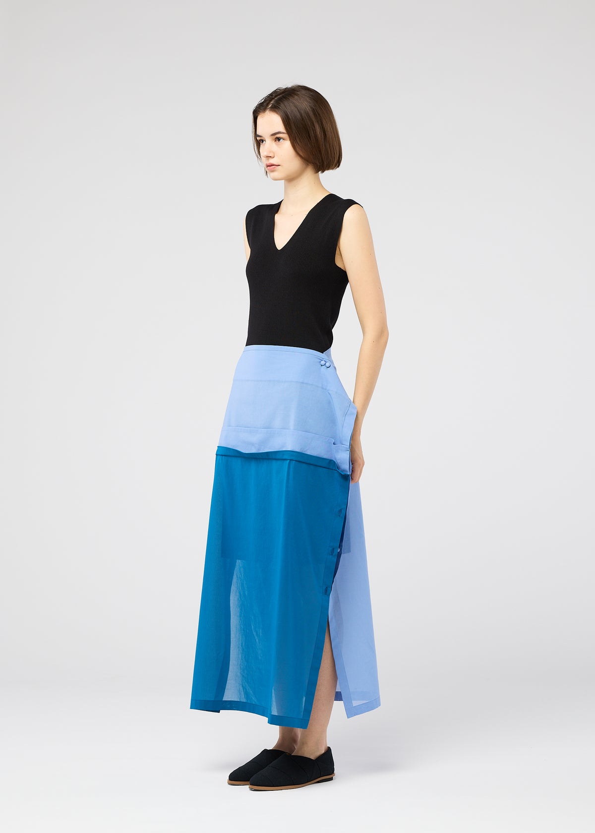 A woman with short brown hair wears the SHEER FOLIO skirt by 132 5. ISSEY MIYAKE—a color-block blue, portfolio-inspired design with side buttons—paired with a sleeveless black top and black slip-on shoes, set against a light grey background. view 8 of 11