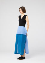 A woman with short brown hair wears the SHEER FOLIO skirt by 132 5. ISSEY MIYAKE—a color-block blue, portfolio-inspired design with side buttons—paired with a sleeveless black top and black slip-on shoes, set against a light grey background.