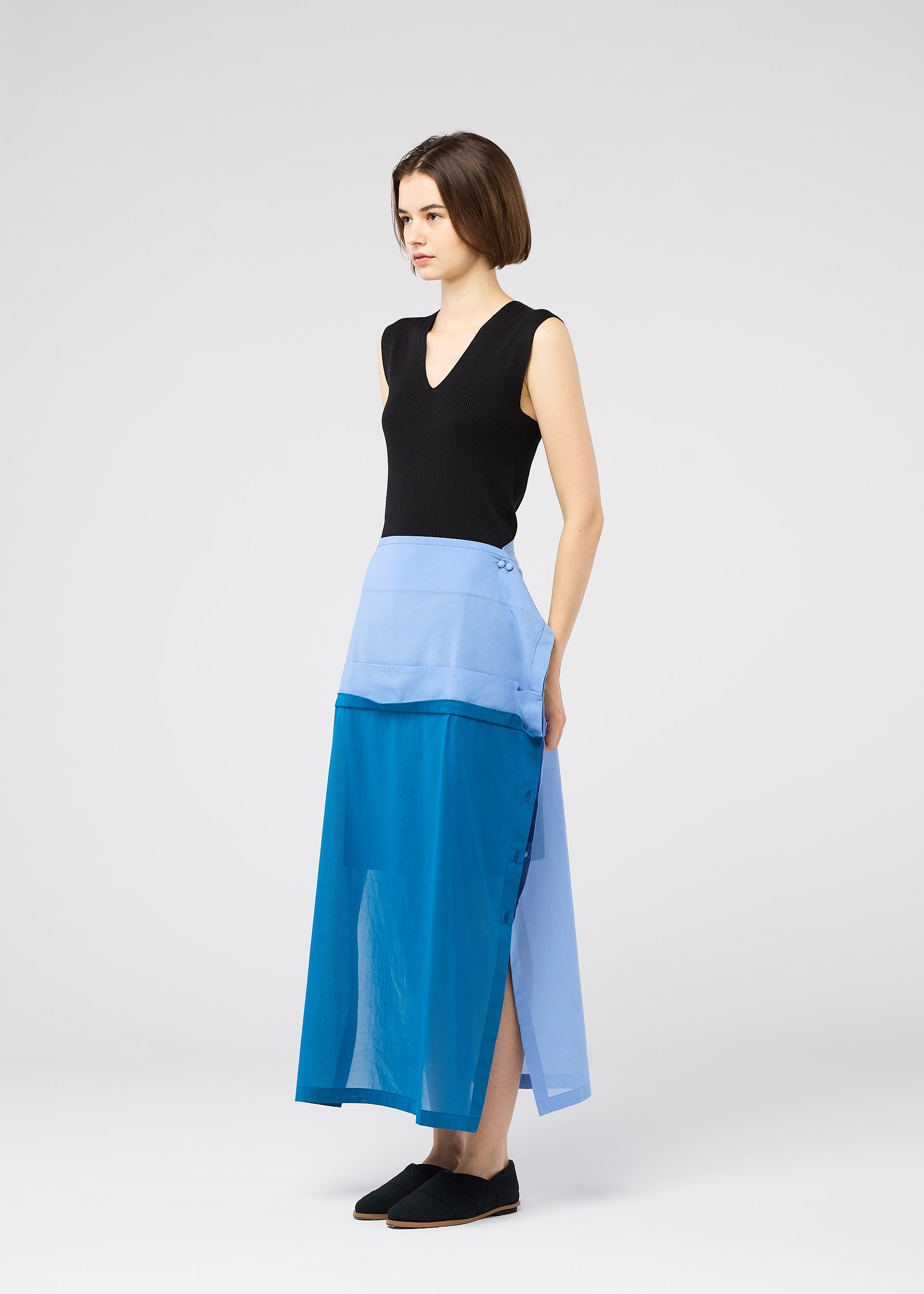 Zoomed view A woman with short brown hair wears the SHEER FOLIO skirt by 132 5. ISSEY MIYAKE—a color-block blue, portfolio-inspired design with side buttons—paired with a sleeveless black top and black slip-on shoes, set against a light grey background.