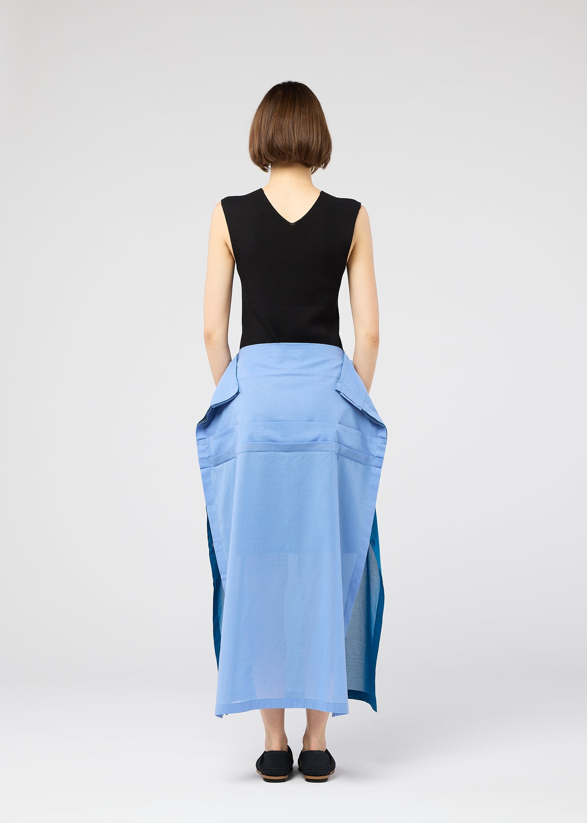 A person with short brown hair wears 132 5. ISSEY MIYAKE’s SHEER FOLIO: a sleeveless black top and blue portfolio-inspired layered skirt with large pockets, paired with black shoes, standing against a plain light background. view 9 of 11
