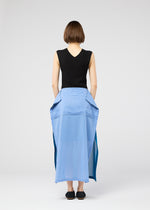 A person with short brown hair wears 132 5. ISSEY MIYAKE’s SHEER FOLIO: a sleeveless black top and blue portfolio-inspired layered skirt with large pockets, paired with black shoes, standing against a plain light background.