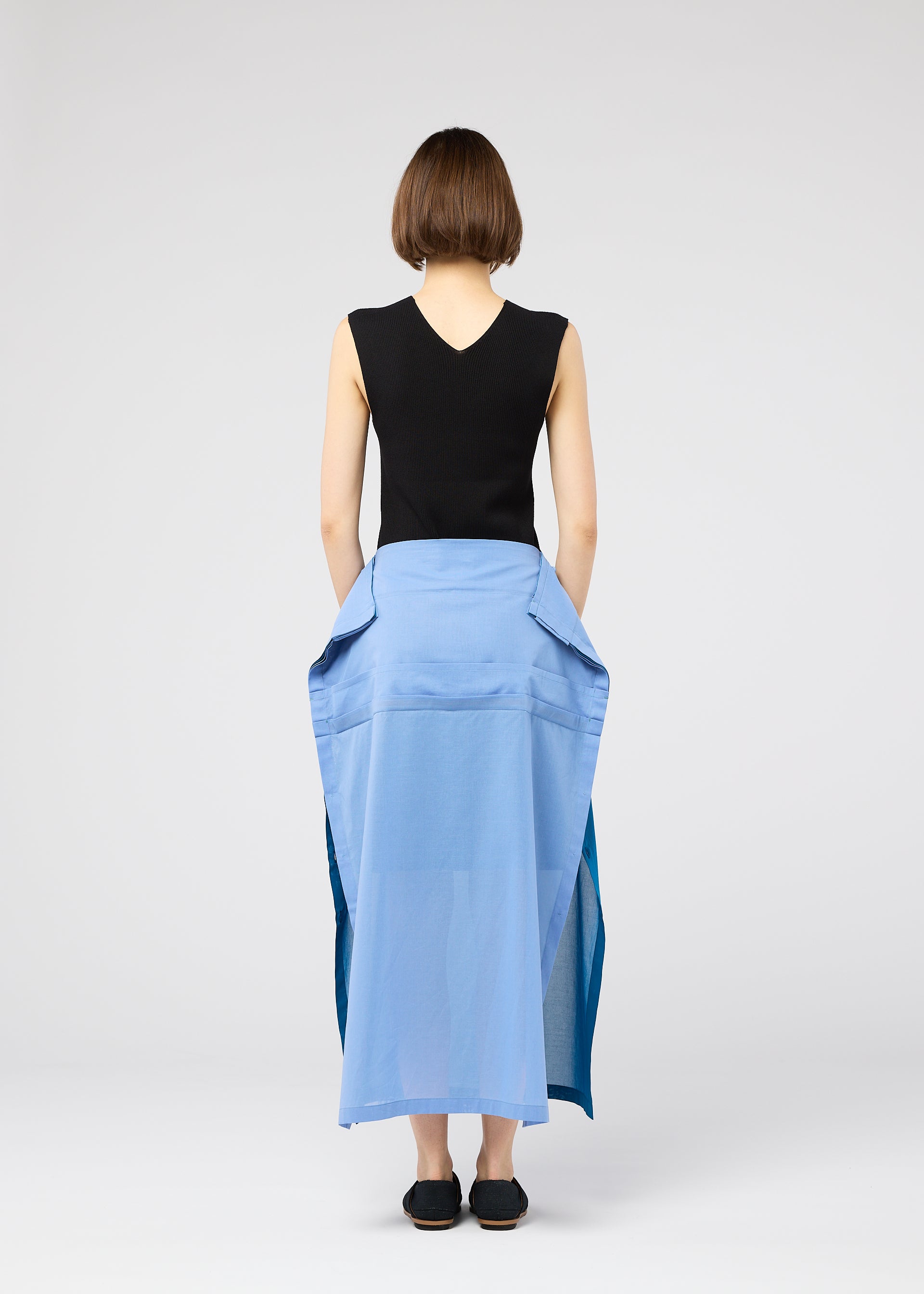 Zoomed view A person with short brown hair wears 132 5. ISSEY MIYAKE’s SHEER FOLIO: a sleeveless black top and blue portfolio-inspired layered skirt with large pockets, paired with black shoes, standing against a plain light background.