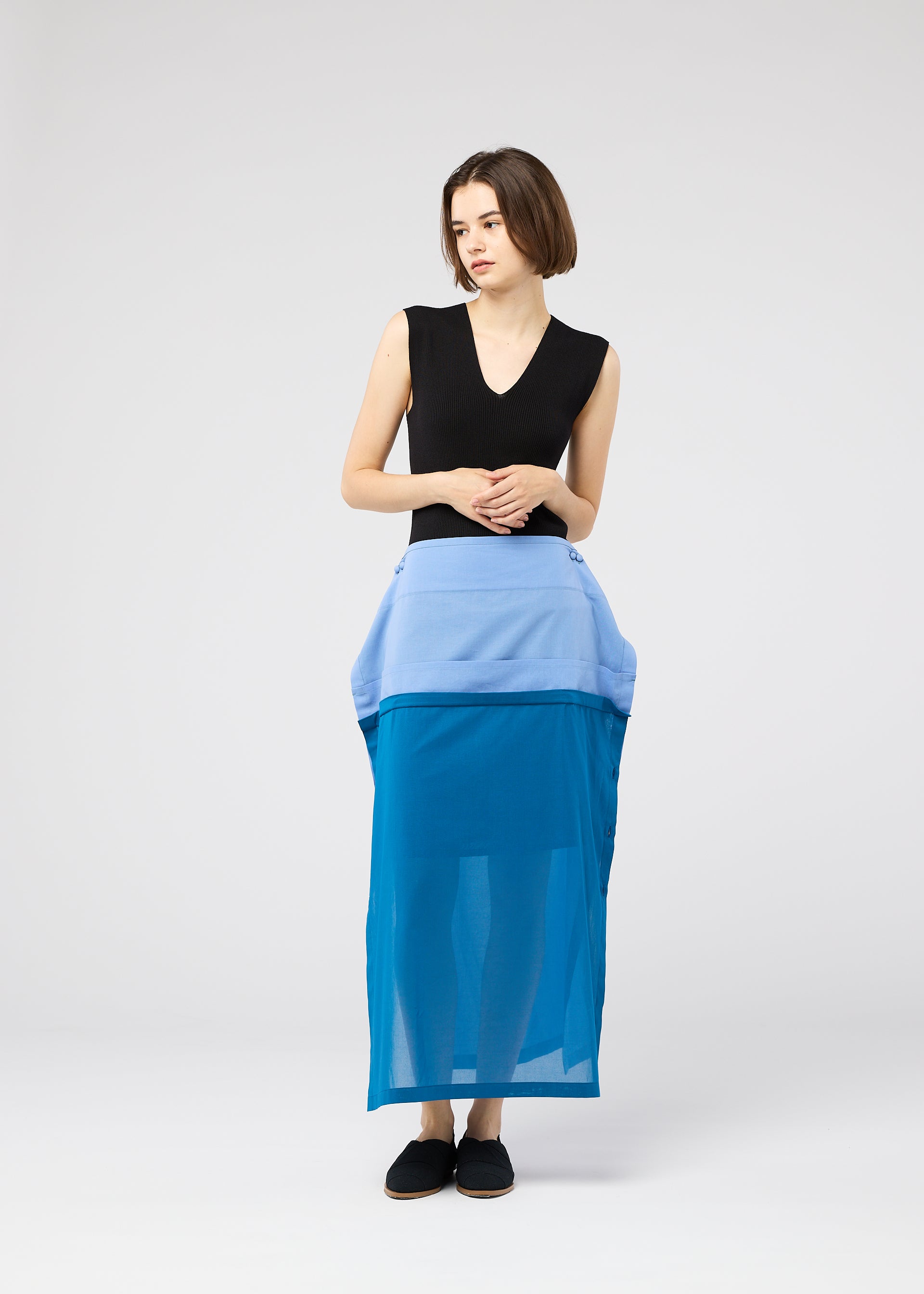 Zoomed view A woman with a short bob wears the SHEER FOLIO layered petticoat skirt by 132 5. ISSEY MIYAKE, paired with a sleeveless black top and black shoes, standing against a plain light gray background.