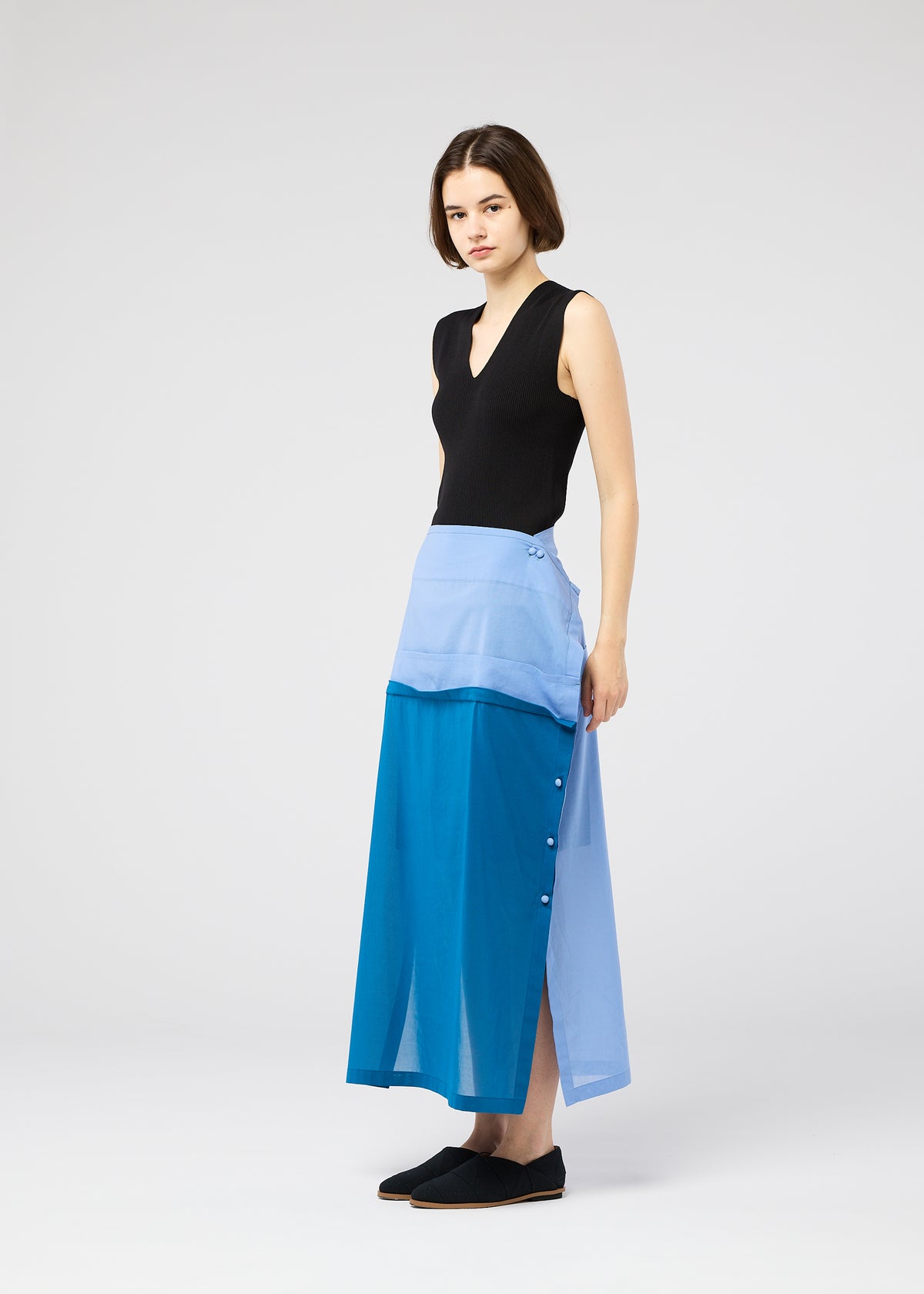A woman with short brown hair wears the 132 5. ISSEY MIYAKE SHEER FOLIO layered skirt with side buttons and a sleeveless black top, standing sideways against a plain white background. view 11 of 11