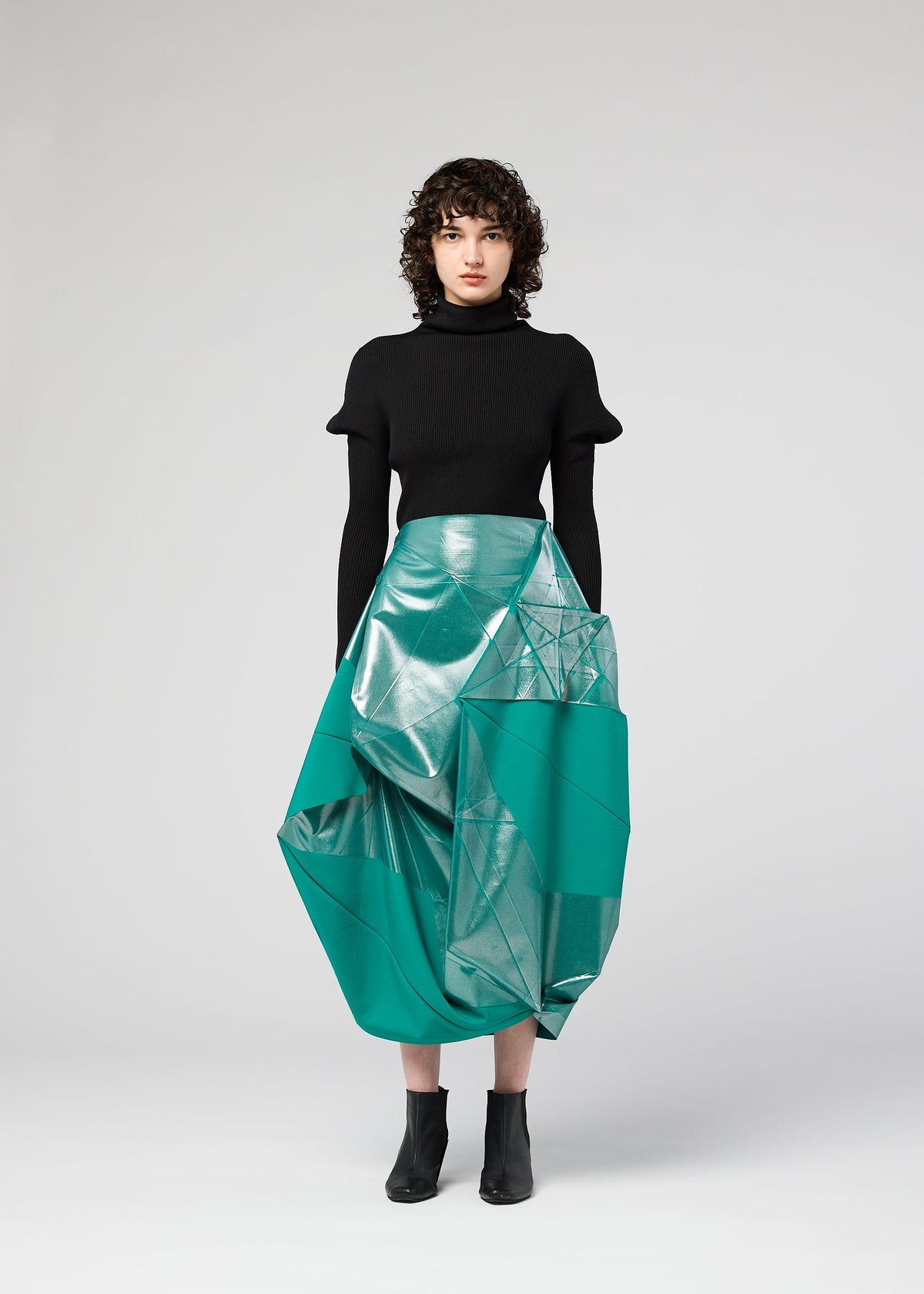 A person with curly hair stands facing forward against a light gray background, wearing 132 5. ISSEY MIYAKE's NO.17 FOIL: a black turtleneck top and a shiny teal skirt with geometric folds, plus black ankle boots. view 7 of 12