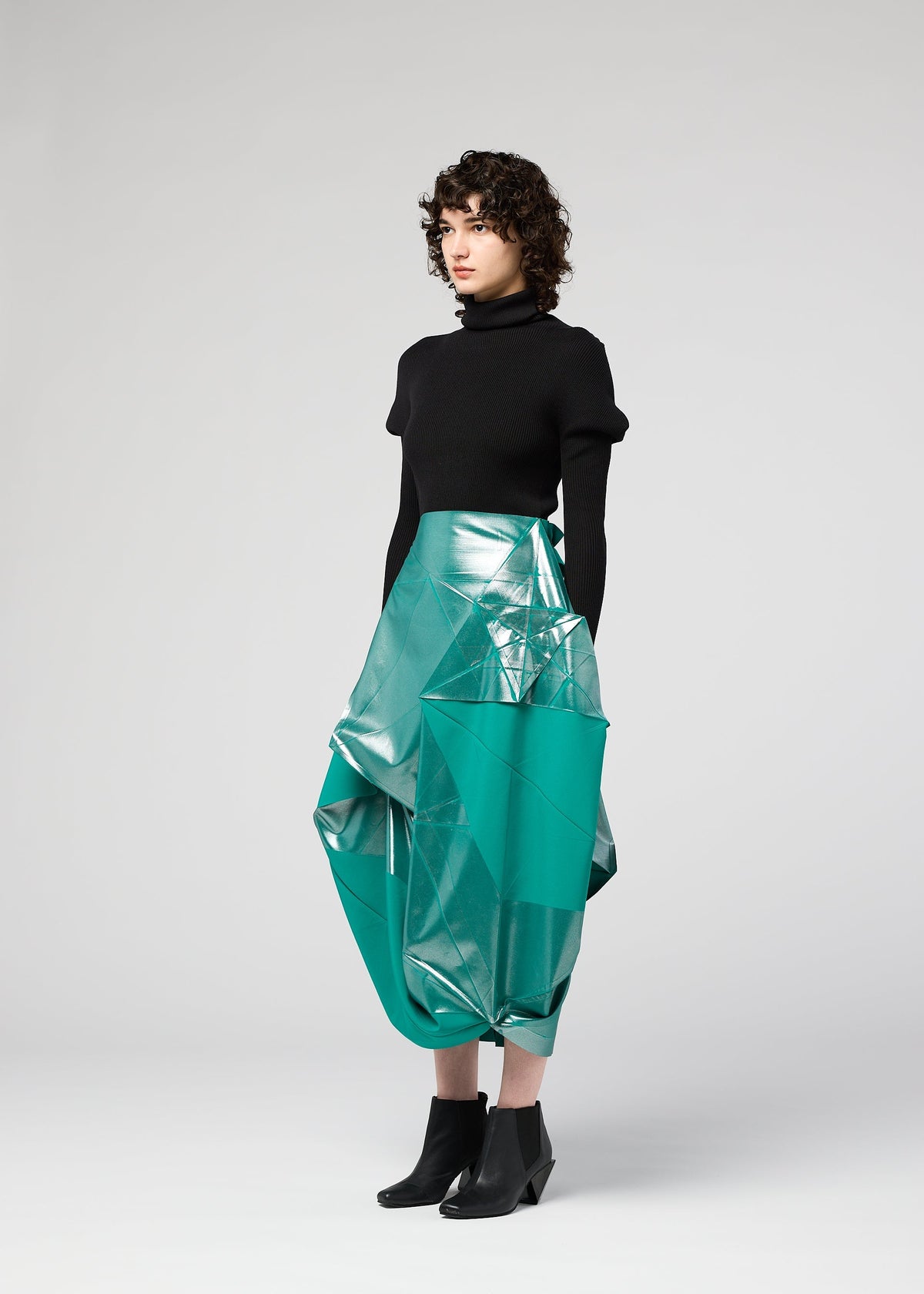 A person with curly hair wears a black turtleneck, the NO.17 FOIL skirt by 132 5. ISSEY MIYAKE—a metallic teal geometric piece with an innovative folding structure—and black ankle boots, standing against a light gray background. view 8 of 12