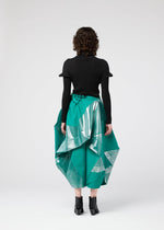 A person with curly hair faces away, wearing the NO.17 FOIL skirt by 132 5. ISSEY MIYAKE—an emerald green piece with a three-dimensional silhouette and metallic silver accents—paired with a black top and ankle boots against a plain light background.