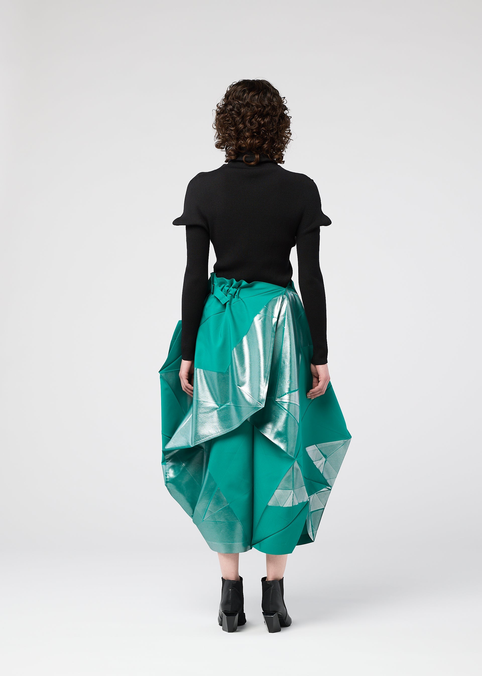 Zoomed view A person with curly hair faces away, wearing the NO.17 FOIL skirt by 132 5. ISSEY MIYAKE—an emerald green piece with a three-dimensional silhouette and metallic silver accents—paired with a black top and ankle boots against a plain light background.
