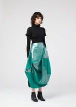A woman with curly brown hair wears the NO.17 FOIL skirt by 132 5. ISSEY MIYAKE—a shiny teal geometric midi with an innovative folding design—paired with a black turtleneck sweater and ankle boots, standing against a light gray background.