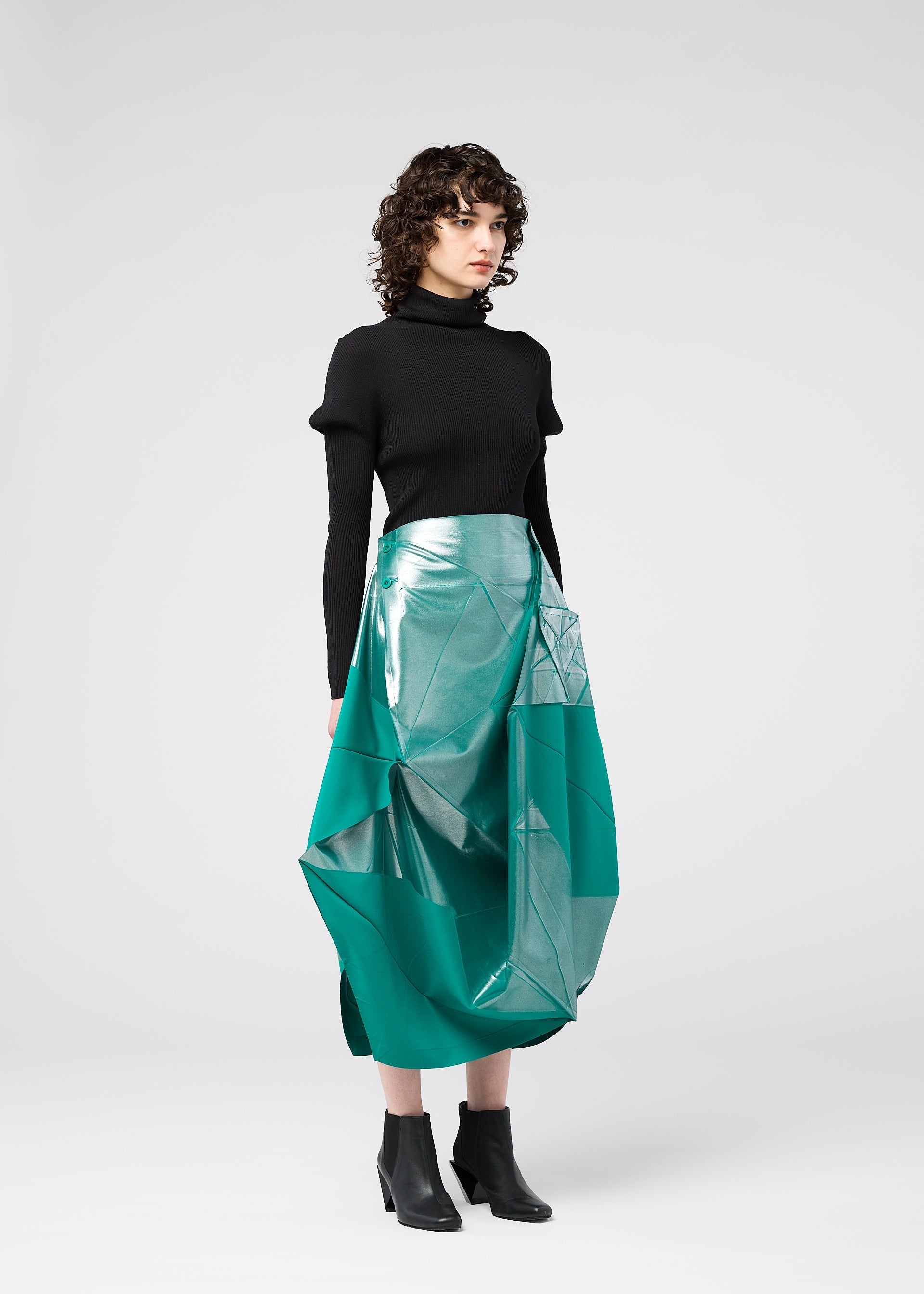 Zoomed view A woman with curly brown hair wears the NO.17 FOIL skirt by 132 5. ISSEY MIYAKE—a shiny teal geometric midi with an innovative folding design—paired with a black turtleneck sweater and ankle boots, standing against a light gray background.