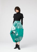 A person with short curly hair wears a fitted black turtleneck, black ankle boots, and the NO.17 FOIL turquoise geometric skirt by 132 5. ISSEY MIYAKE, standing against a plain light background.