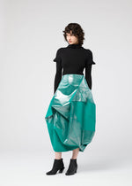 A person with short, curly hair wears a black turtleneck, black ankle boots, and the NO.17 FOIL skirt by 132 5. ISSEY MIYAKE—a shiny turquoise geometric design—standing against a plain light background.