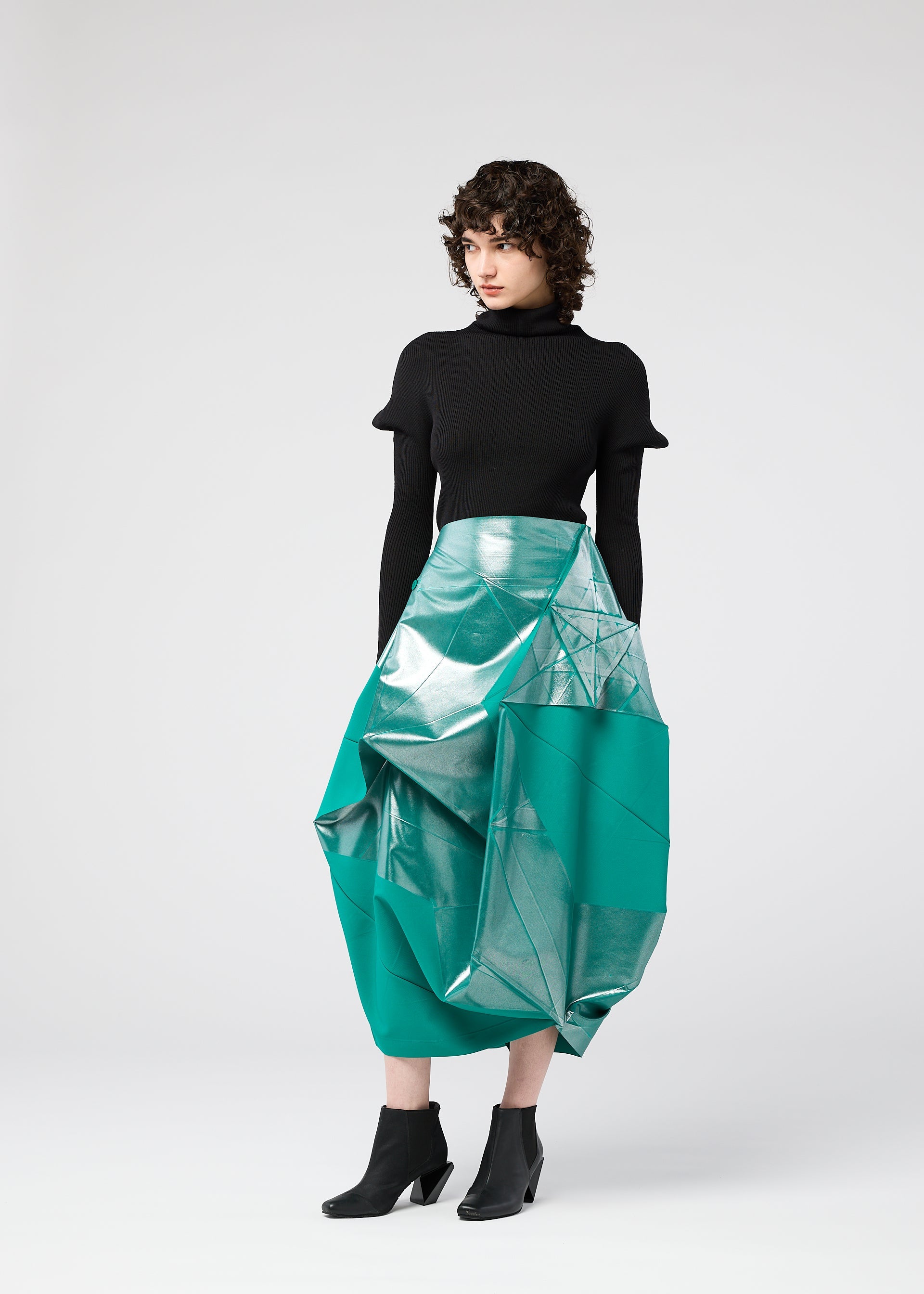 Zoomed view A person with short, curly hair wears a black turtleneck, black ankle boots, and the NO.17 FOIL skirt by 132 5. ISSEY MIYAKE—a shiny turquoise geometric design—standing against a plain light background.