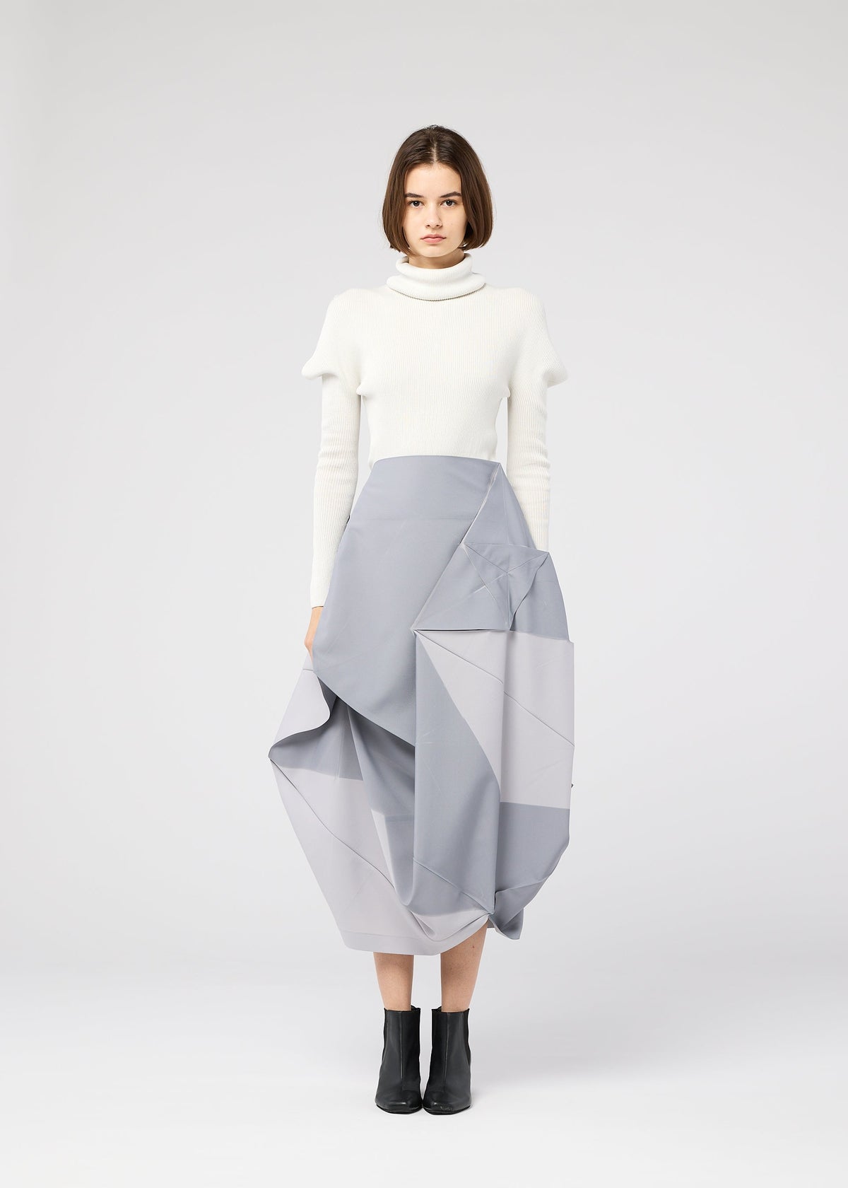 A woman with straight brown hair and a neutral expression wears the 132 5. ISSEY MIYAKE NO.17 PRINT light gray folding structure skirt, a white turtleneck sweater, and black ankle boots against a plain background. view 7 of 11