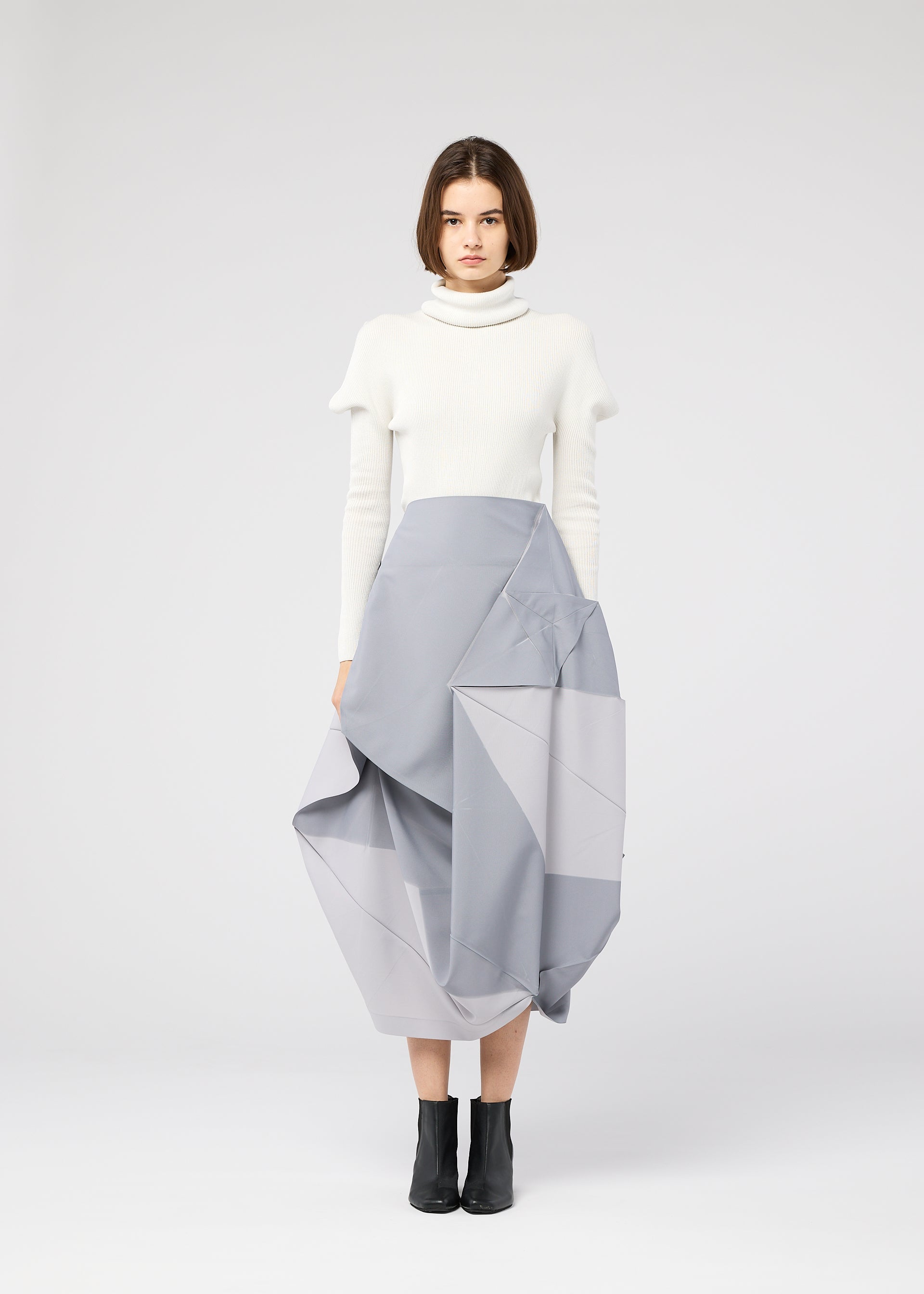 Zoomed view A woman with straight brown hair and a neutral expression wears the 132 5. ISSEY MIYAKE NO.17 PRINT light gray folding structure skirt, a white turtleneck sweater, and black ankle boots against a plain background.