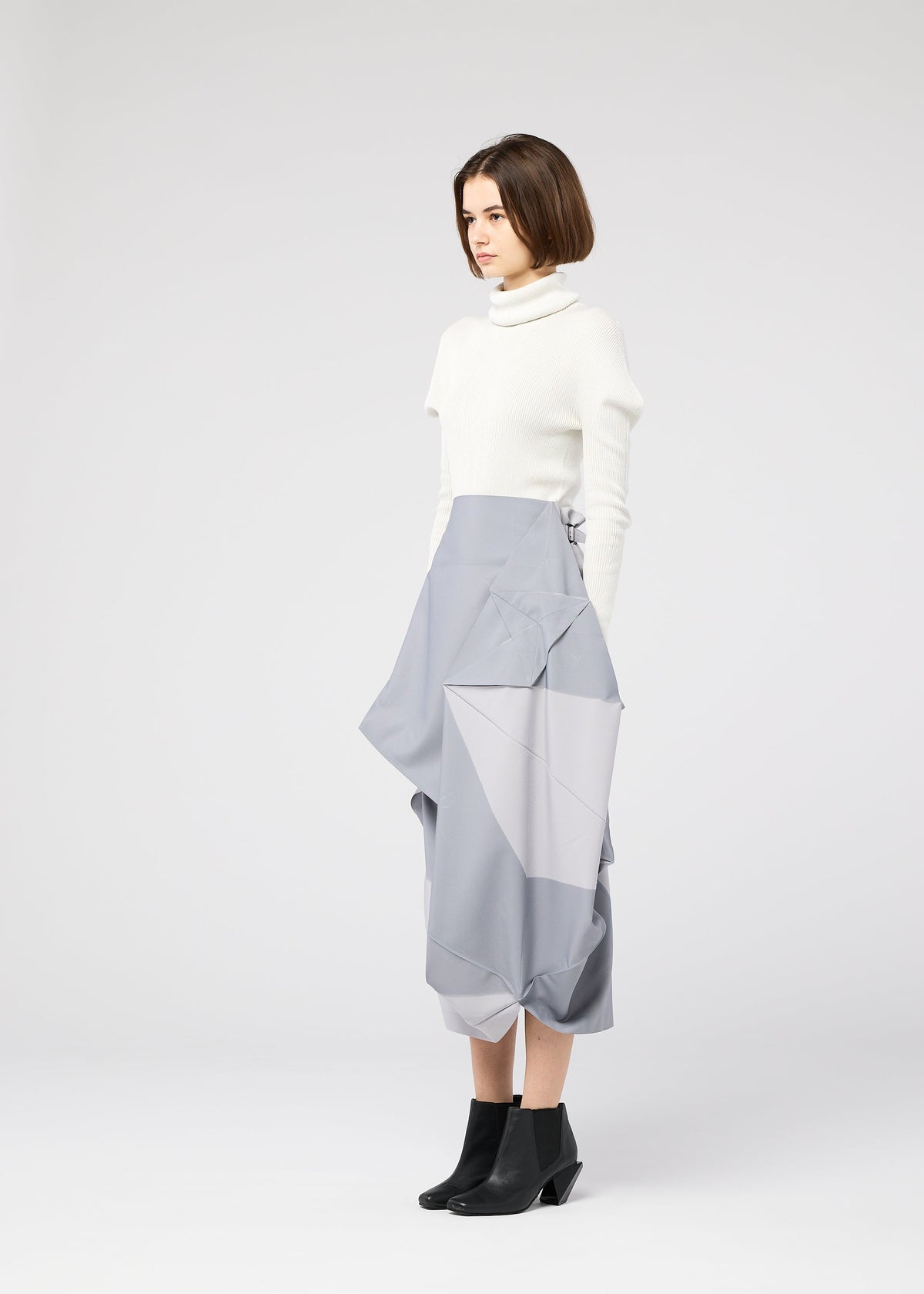 A woman with short brown hair wears the NO.17 PRINT high-waisted geometric fold skirt in light gray by 132 5. ISSEY MIYAKE, paired with a white turtleneck and black ankle boots, standing against a plain light gray background. view 8 of 11