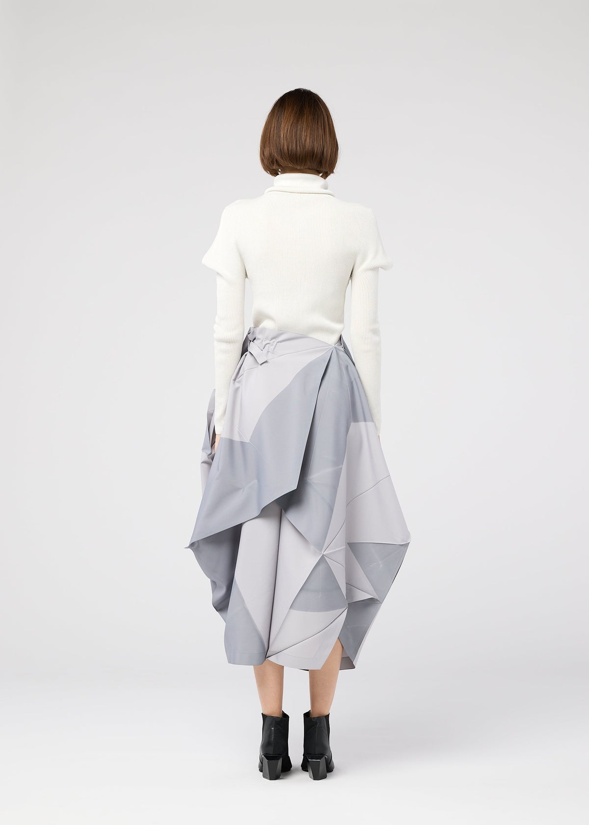 A person wearing the NO.17 PRINT skirt by 132 5. ISSEY MIYAKE, a white turtleneck, black ankle boots, and sporting short brown hair stands facing away against a plain white background. The skirt features geometric, voluminous folds. view 9 of 11