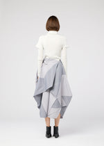 A person wearing the NO.17 PRINT skirt by 132 5. ISSEY MIYAKE, a white turtleneck, black ankle boots, and sporting short brown hair stands facing away against a plain white background. The skirt features geometric, voluminous folds.