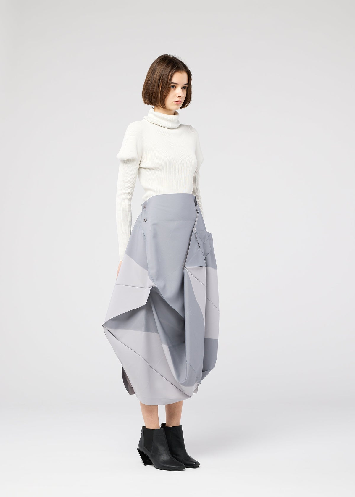 A woman with a bob haircut stands in profile, wearing the NO.17 PRINT skirt by 132 5. ISSEY MIYAKE, paired with a white turtleneck sweater and black ankle boots, set against a plain light gray background. view 10 of 11