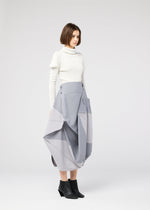A woman with a bob haircut stands in profile, wearing the NO.17 PRINT skirt by 132 5. ISSEY MIYAKE, paired with a white turtleneck sweater and black ankle boots, set against a plain light gray background.