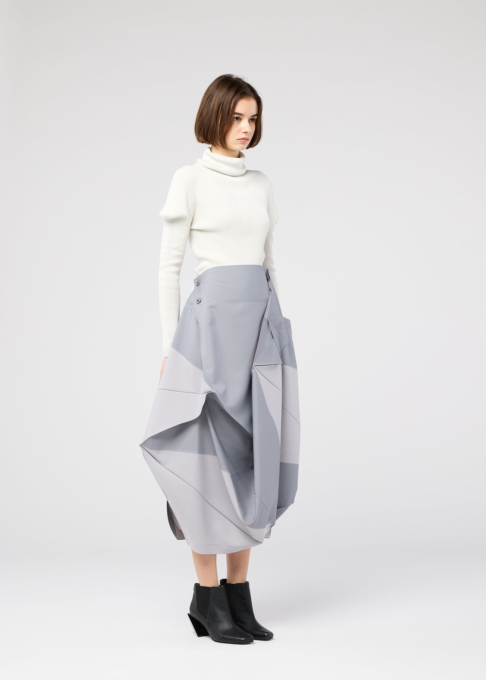 Zoomed view A woman with a bob haircut stands in profile, wearing the NO.17 PRINT skirt by 132 5. ISSEY MIYAKE, paired with a white turtleneck sweater and black ankle boots, set against a plain light gray background.