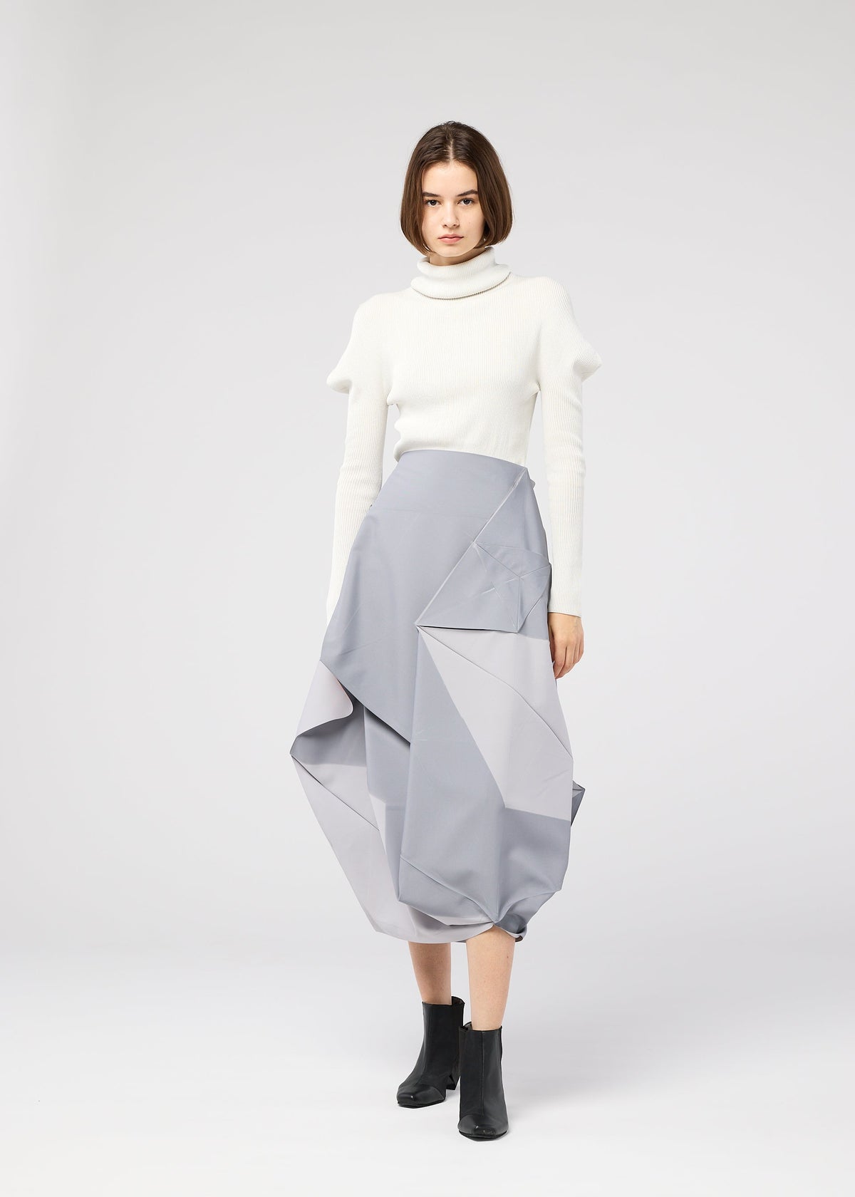 A woman with a bob haircut wears the NO.17 PRINT skirt by 132 5. ISSEY MIYAKE in sculptural light gray, styled with a white turtleneck and black ankle boots, standing against a plain light background. view 11 of 11