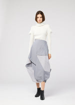 A woman with a bob haircut wears the NO.17 PRINT skirt by 132 5. ISSEY MIYAKE in sculptural light gray, styled with a white turtleneck and black ankle boots, standing against a plain light background.