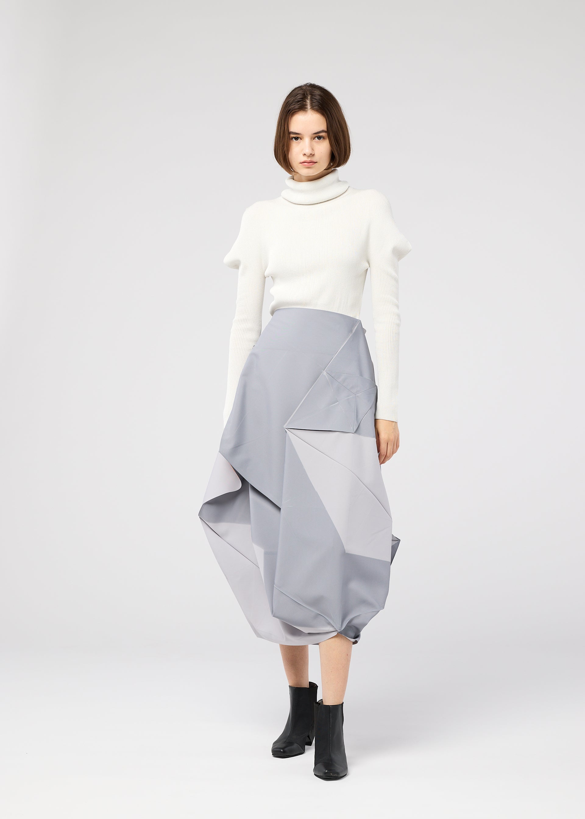 Zoomed view A woman with a bob haircut wears the NO.17 PRINT skirt by 132 5. ISSEY MIYAKE in sculptural light gray, styled with a white turtleneck and black ankle boots, standing against a plain light background.