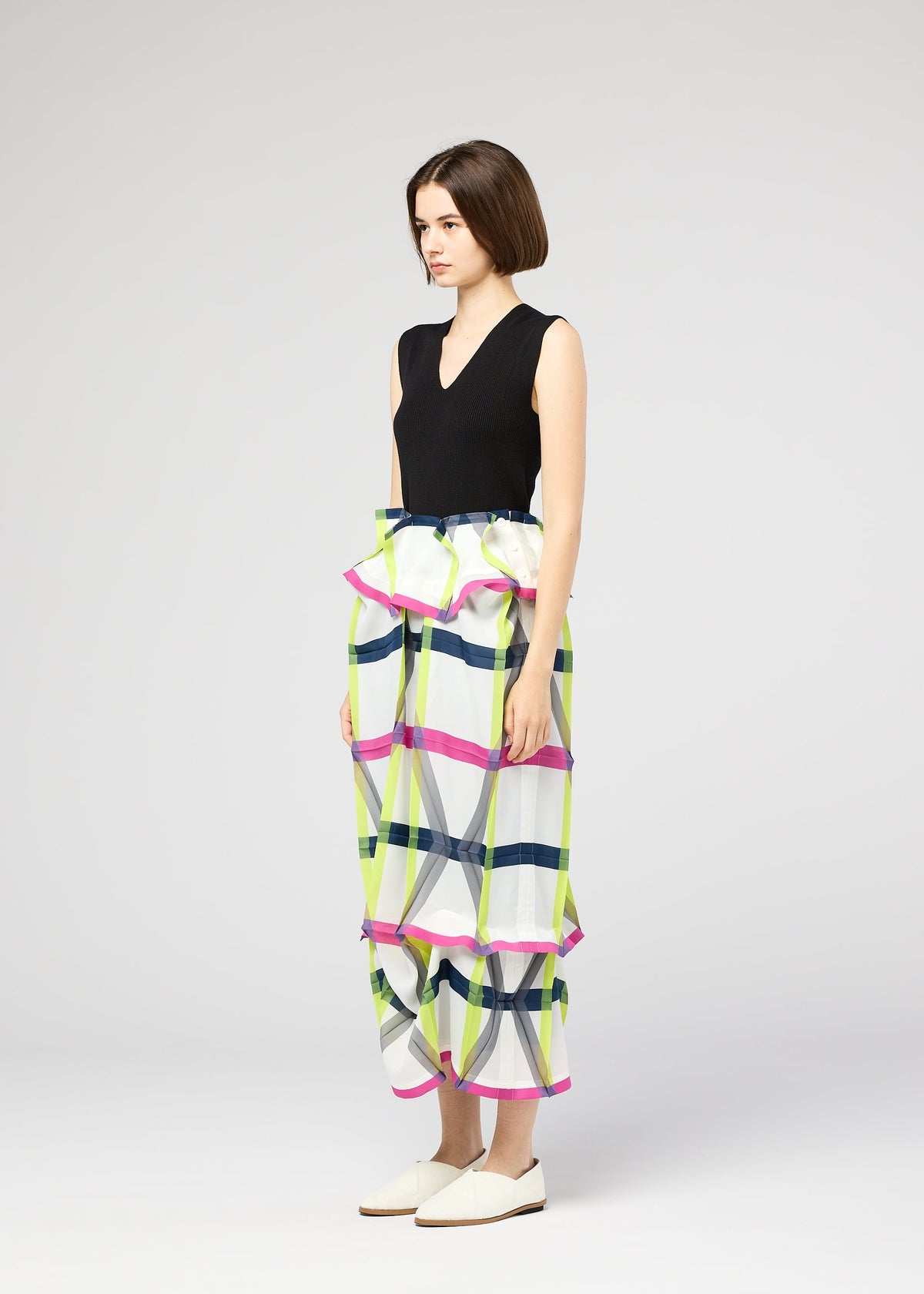 A woman with a short bob wears the NO.6 TRANSLUCENT skirt by 132 5. ISSEY MIYAKE—a high-waisted, colorful plaid piece with ruffles, pleats, and unique folds—paired with a sleeveless black top and white slip-on shoes, standing sideways. view 10 of 13