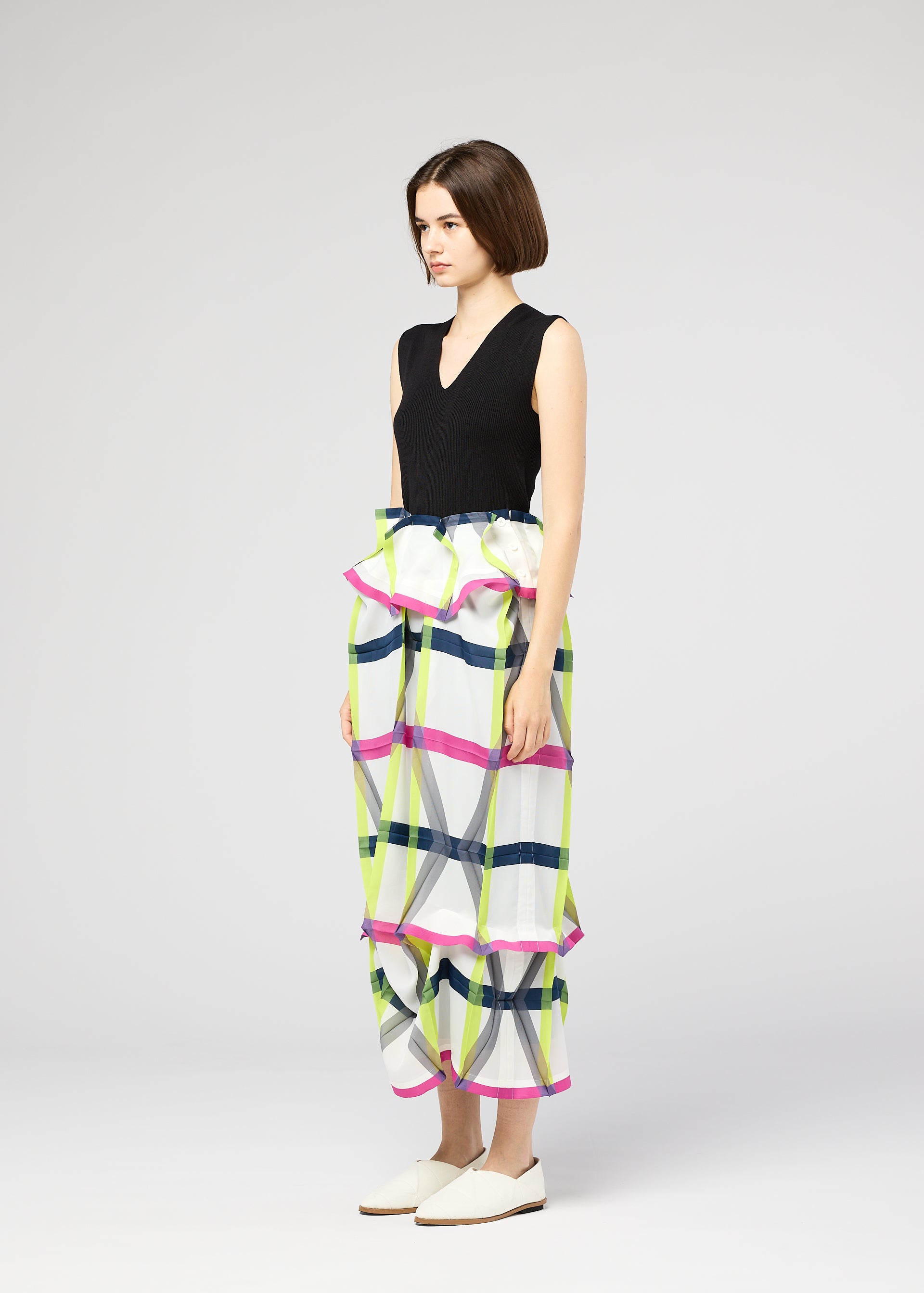 Zoomed view A woman with a short bob wears the NO.6 TRANSLUCENT skirt by 132 5. ISSEY MIYAKE—a high-waisted, colorful plaid piece with ruffles, pleats, and unique folds—paired with a sleeveless black top and white slip-on shoes, standing sideways.