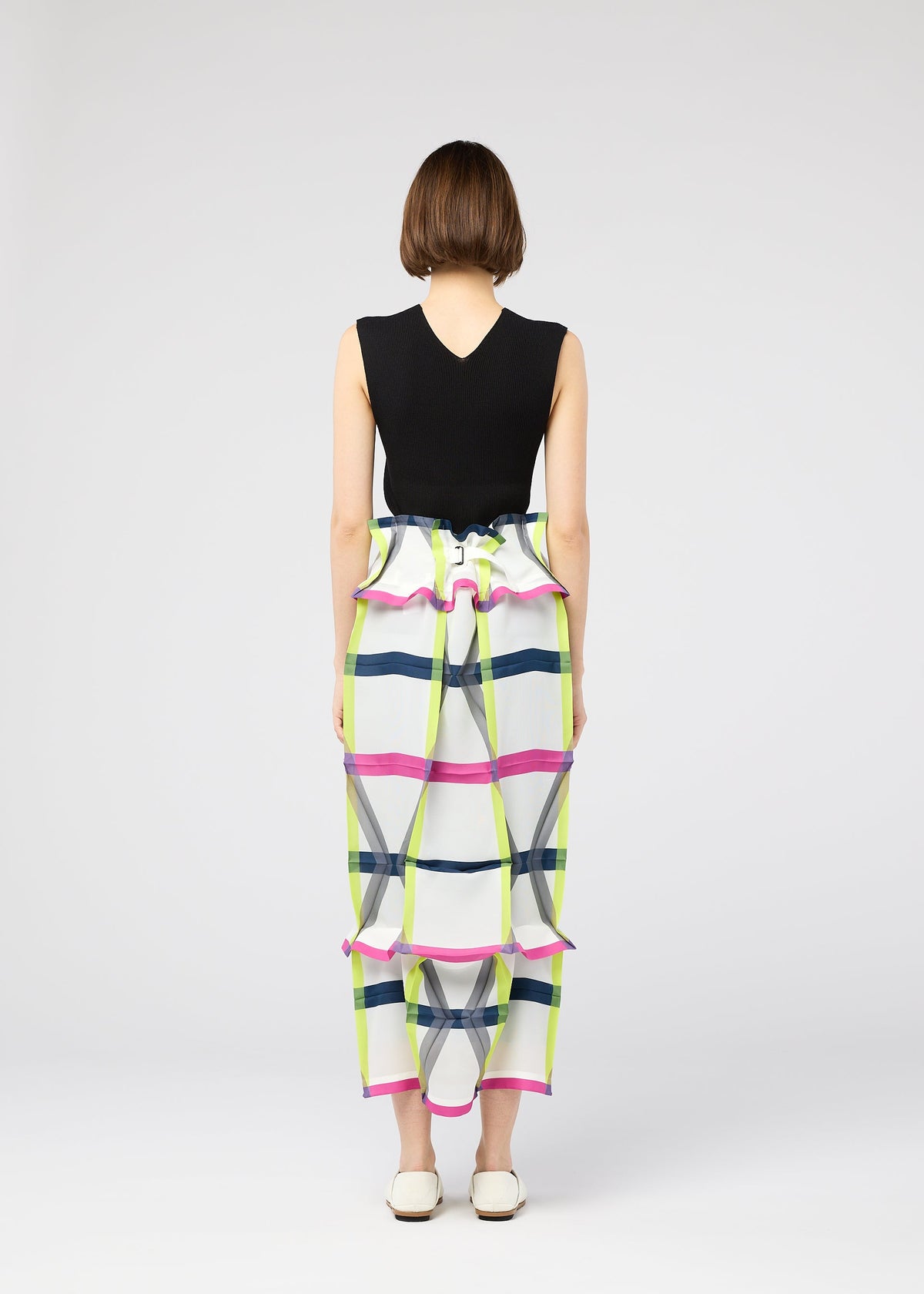 A woman with short brown hair wears the 132 5. ISSEY MIYAKE NO.6 TRANSLUCENT: a sleeveless black top and a long, colorful plaid skirt with ruffled accents and elegant folds, paired with white flats against a light background. view 11 of 13
