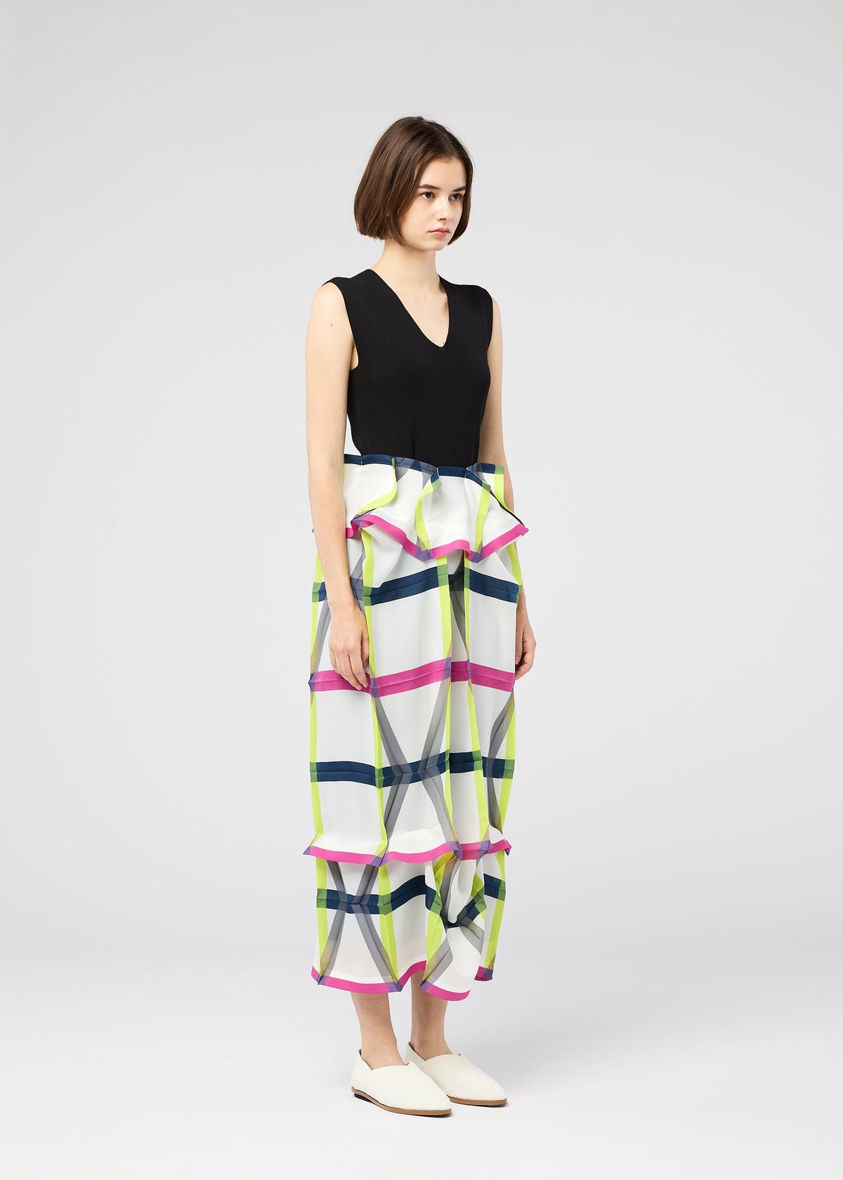 A woman with a neutral expression wears the NO.6 TRANSLUCENT top and wide-leg pants by 132 5. ISSEY MIYAKE, featuring colorful geometric patterns and ruffle details, paired with white flats against a plain light background. view 12 of 13
