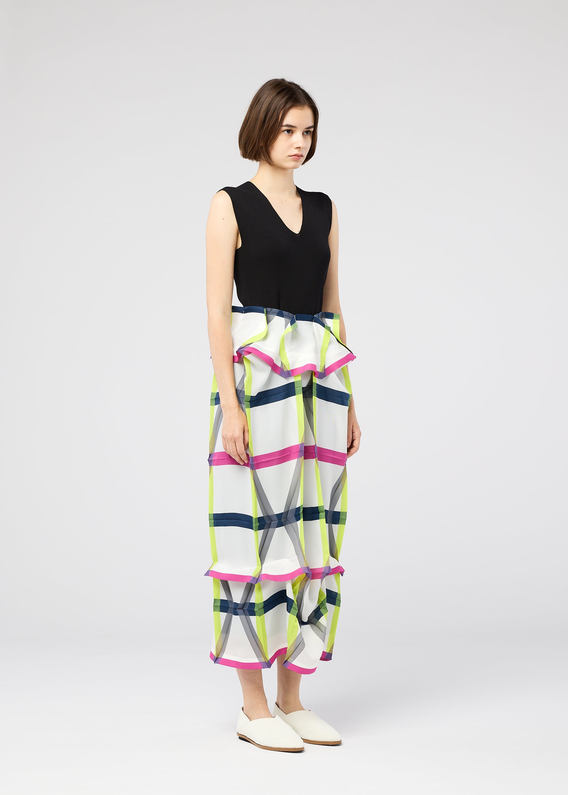 Zoomed view A woman with a neutral expression wears the NO.6 TRANSLUCENT top and wide-leg pants by 132 5. ISSEY MIYAKE, featuring colorful geometric patterns and ruffle details, paired with white flats against a plain light background.